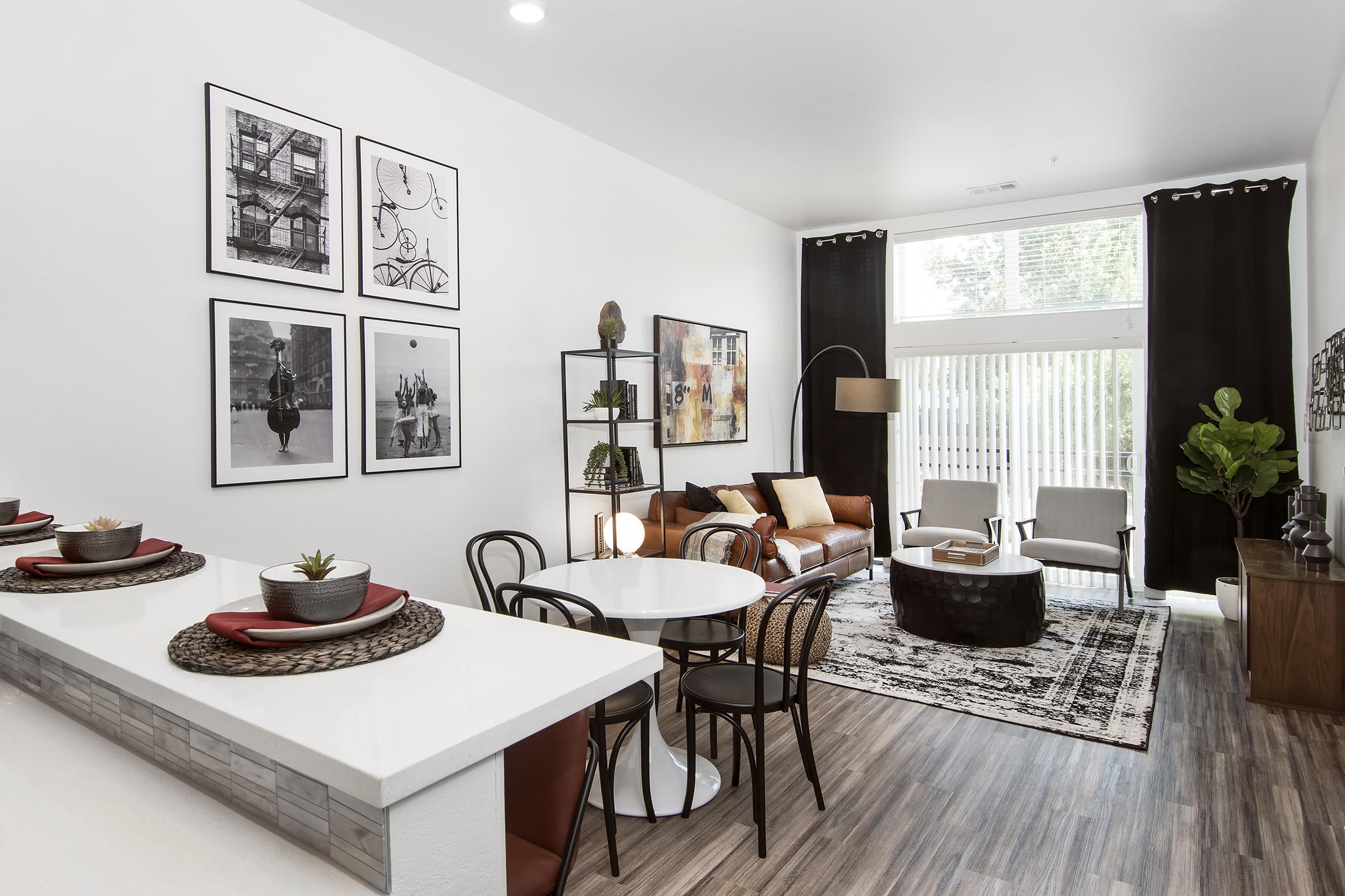 A modern living space featuring a cozy seating area with a brown sofa, two light chairs, a round dining table with black chairs, and decorative wall art. Large windows allow natural light, and there are plants and stylish decor elements throughout the room, creating a warm and inviting atmosphere.