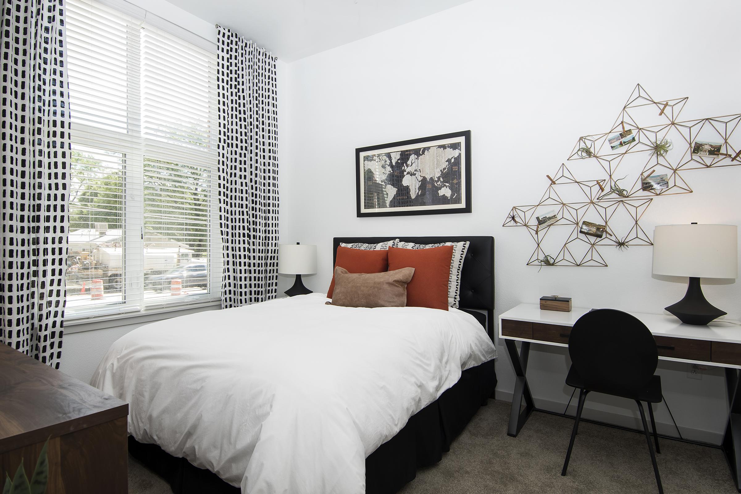 A cozy, modern bedroom featuring a black and white color scheme. The room has a bed with white bedding and orange accent pillows, a stylish wall art piece, a small desk with a chair, and large windows covered with patterned curtains, allowing natural light to fill the space.