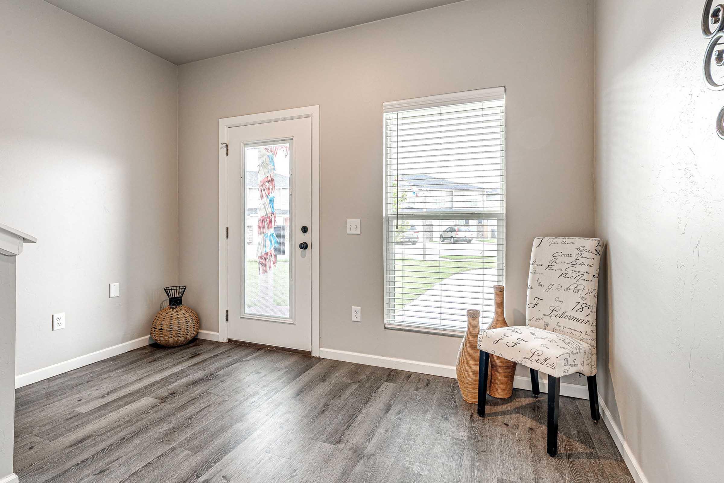 An entryway featuring a door with glass panels, allowing natural light, and a window with blinds. There is a decorative chair with a patterned fabric, a tall woven basket, and a wooden vase. The floor has a wood-like finish, creating a warm and inviting atmosphere.