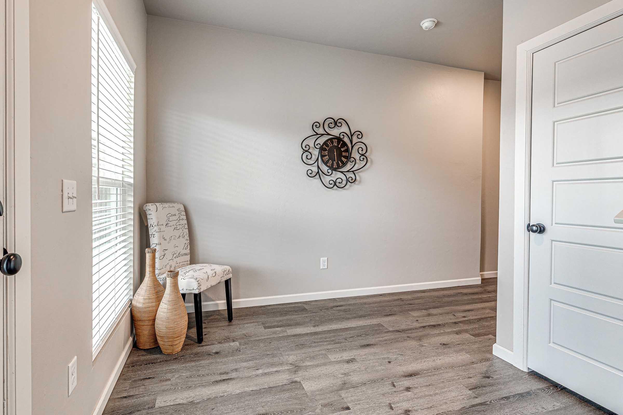 A simple interior space featuring a chair with an elegant design, two decorative vases on the floor, a wall clock, and natural light streaming through a window. The walls are painted in light gray, and the flooring is a smooth, dark wood. A door is visible on the right side.