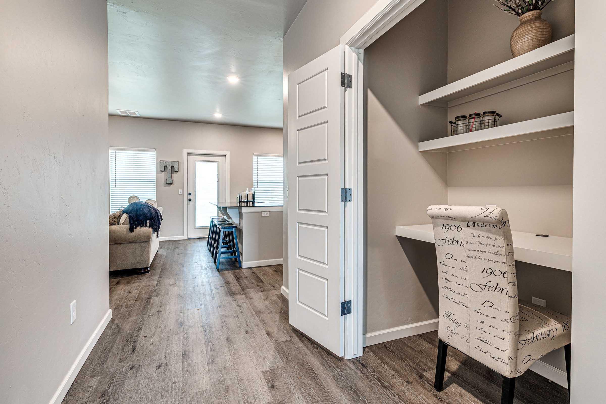A modern hallway featuring light gray walls and wood flooring. On the left, a small nook with a chair and shelves. At the end of the hallway, a living area with a couch and a kitchen counter is visible. Natural light comes through windows, creating a bright and inviting atmosphere.