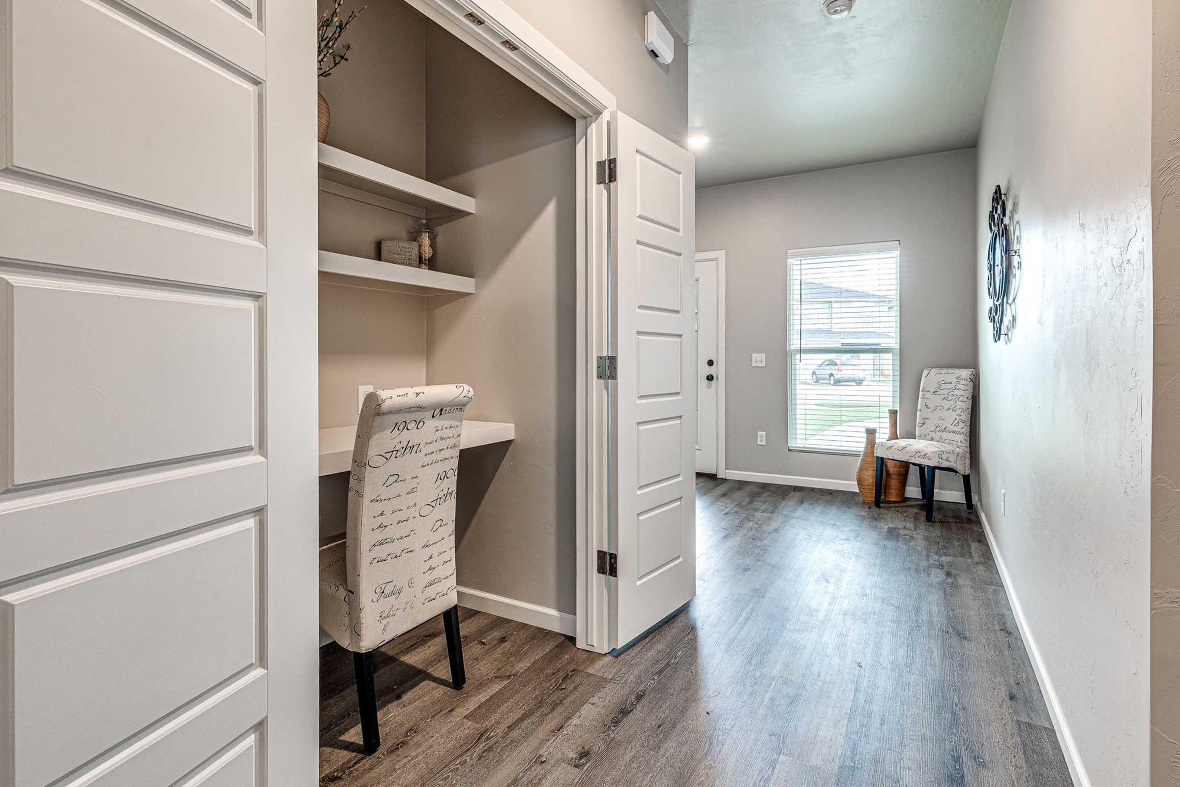 A modern hallway features a desk with a patterned chair, open shelves on the wall, and a cozy seating area near a window. The walls are painted in neutral tones, and the flooring is dark wood. A decorative mirror adds style, while natural light brightens the space.