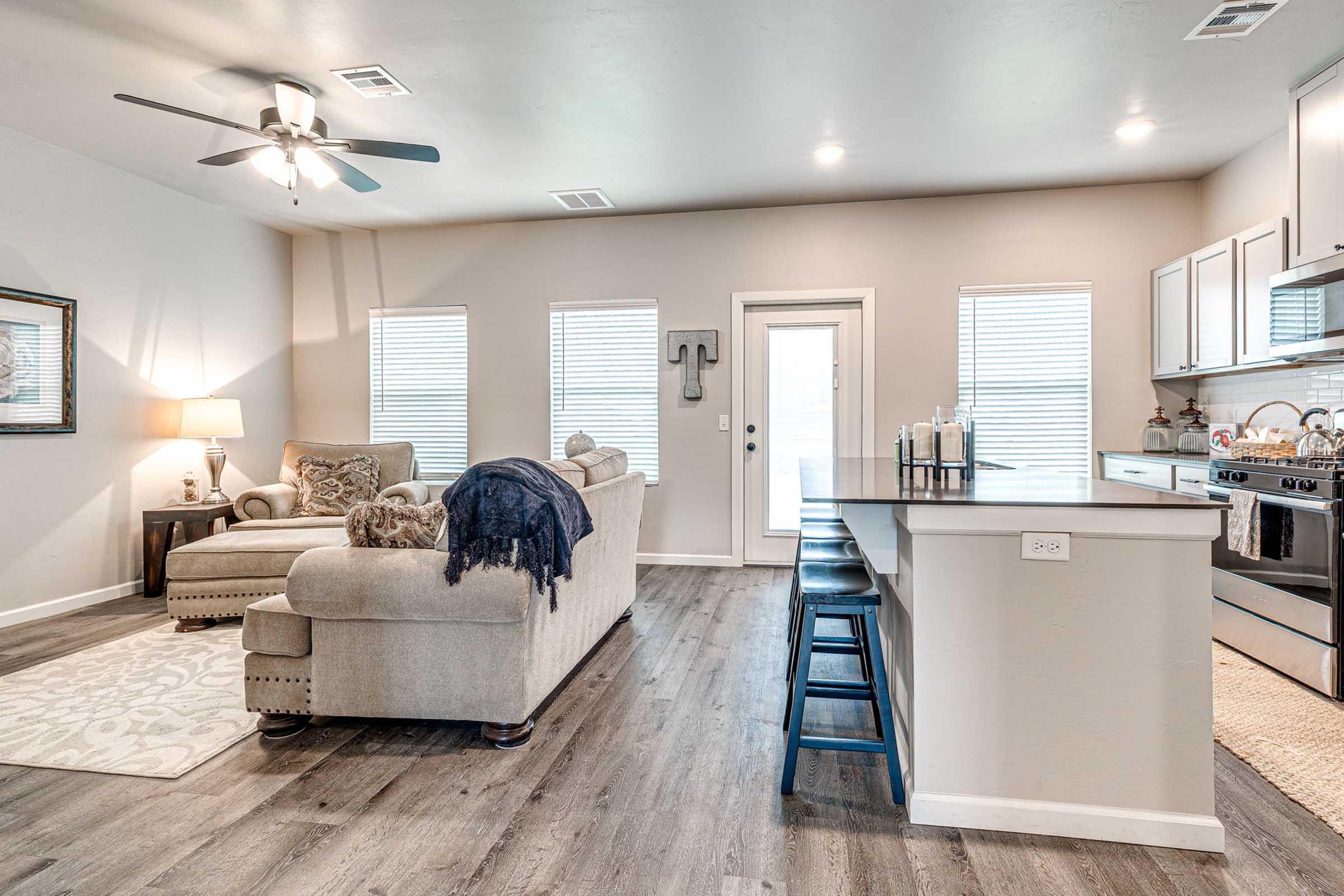 Modern living room and kitchen area featuring a cozy sectional couch with decorative pillows, a coffee table, and a ceiling fan. The open space includes a dining bar with stools, light-colored cabinetry, and large windows providing natural light. Wood-look flooring adds warmth to the contemporary design.