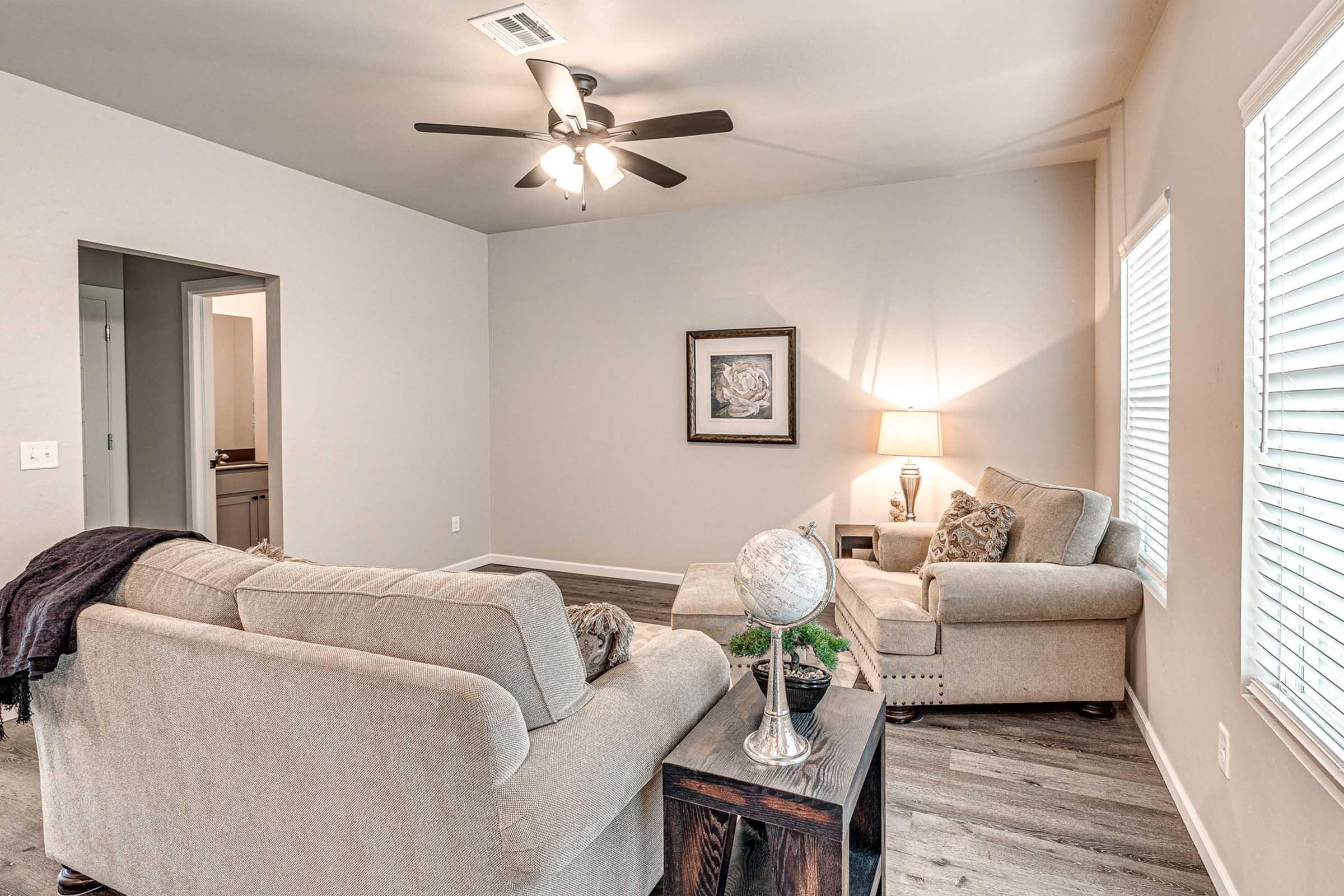 A cozy living room with light-colored walls and a ceiling fan. Two beige sofas are positioned around a small table featuring a globe. A lamp on the table provides soft lighting, and a framed artwork hangs on the wall. Natural light streams in through window blinds.