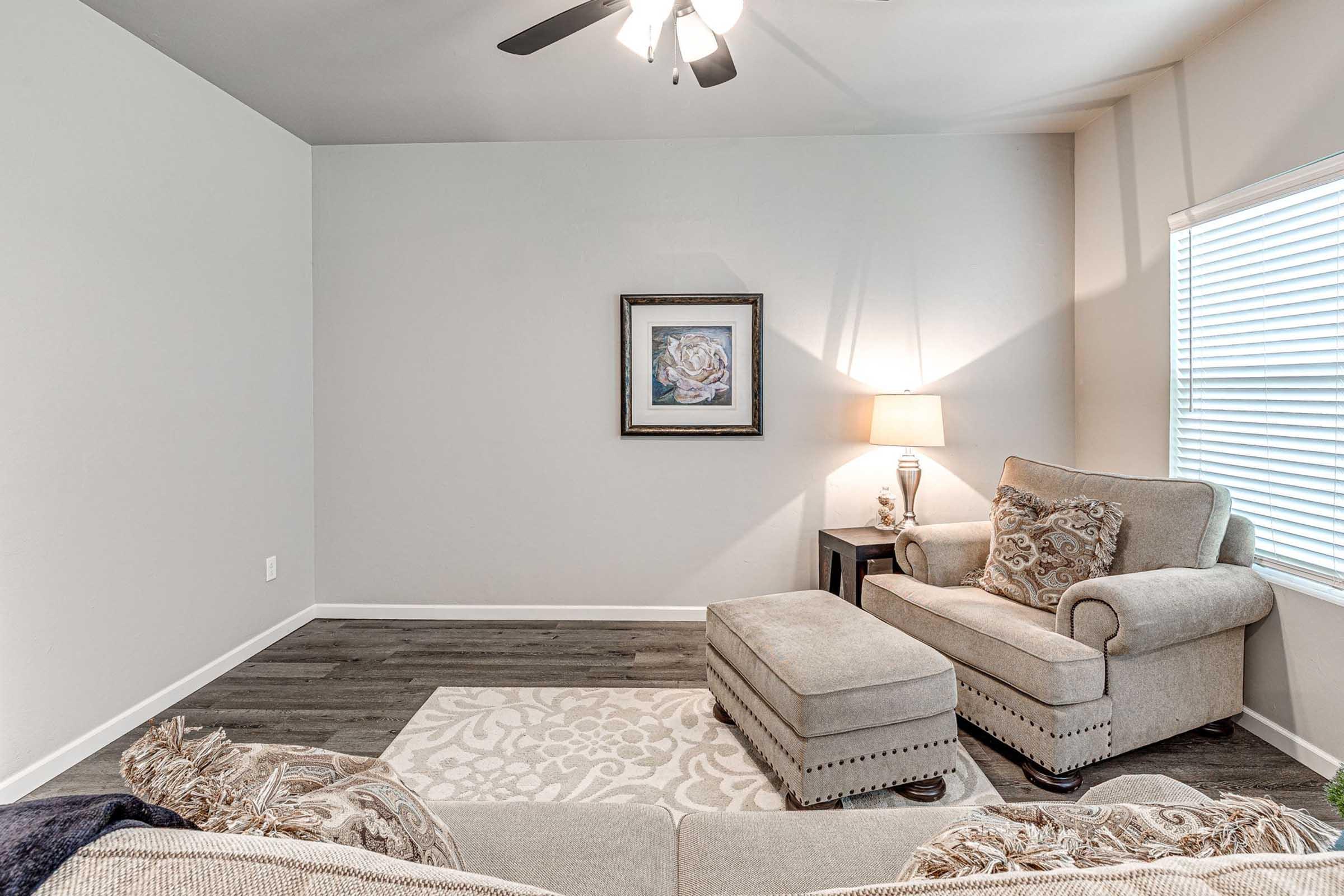 A cozy living room featuring a light-colored sofa with decorative pillows, an ottoman, and a side table with a lamp. The walls are painted in soft tones, and a piece of framed artwork hangs above the sofa. There's a window with blinds allowing natural light to brighten the space.