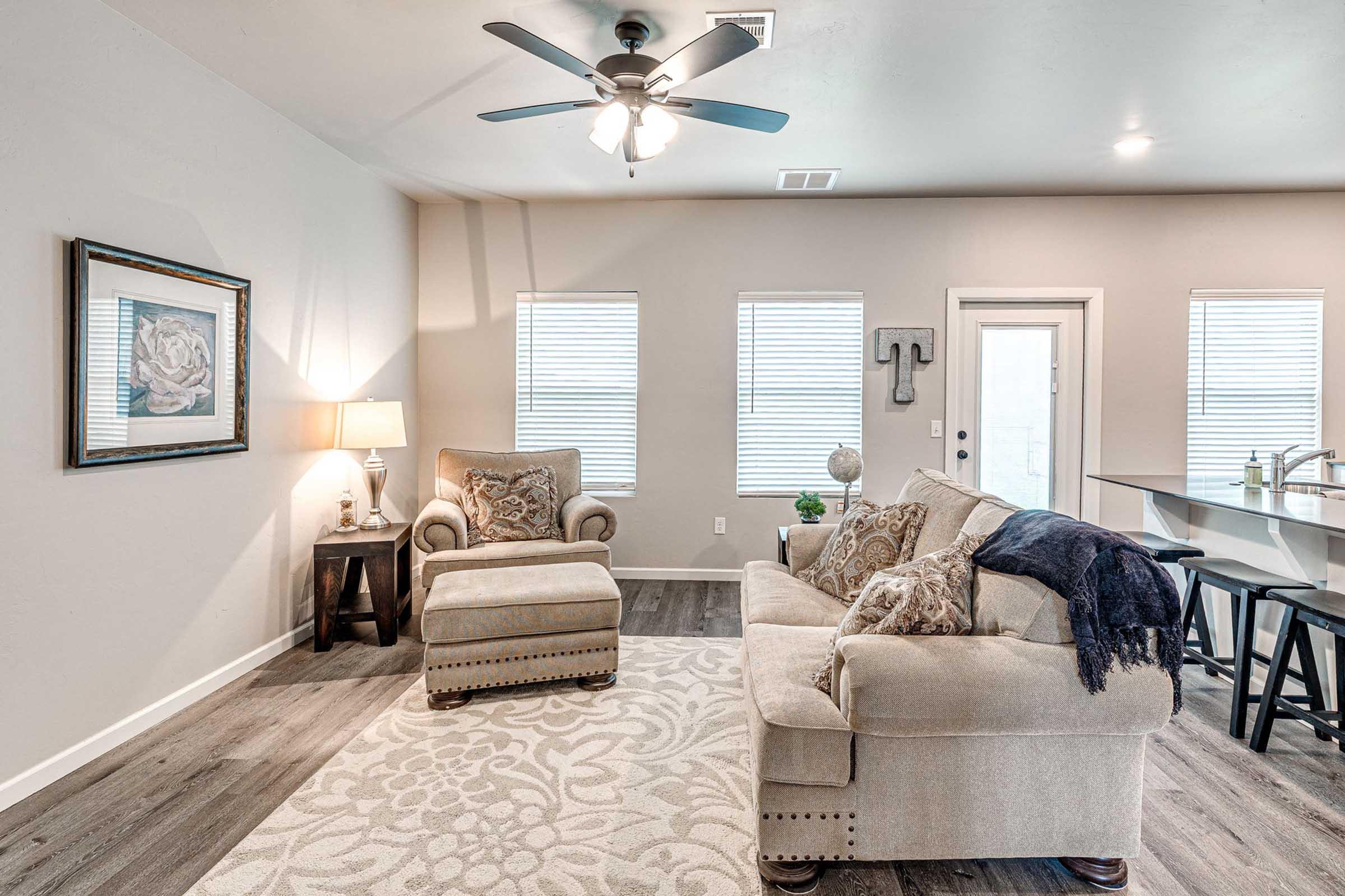 Cozy living room featuring a light-colored sofa with decorative pillows, an armchair, a ceiling fan, and a small side table with a lamp. The room has large windows with white blinds, a light-colored area rug, and a bar-style kitchen counter in the background.