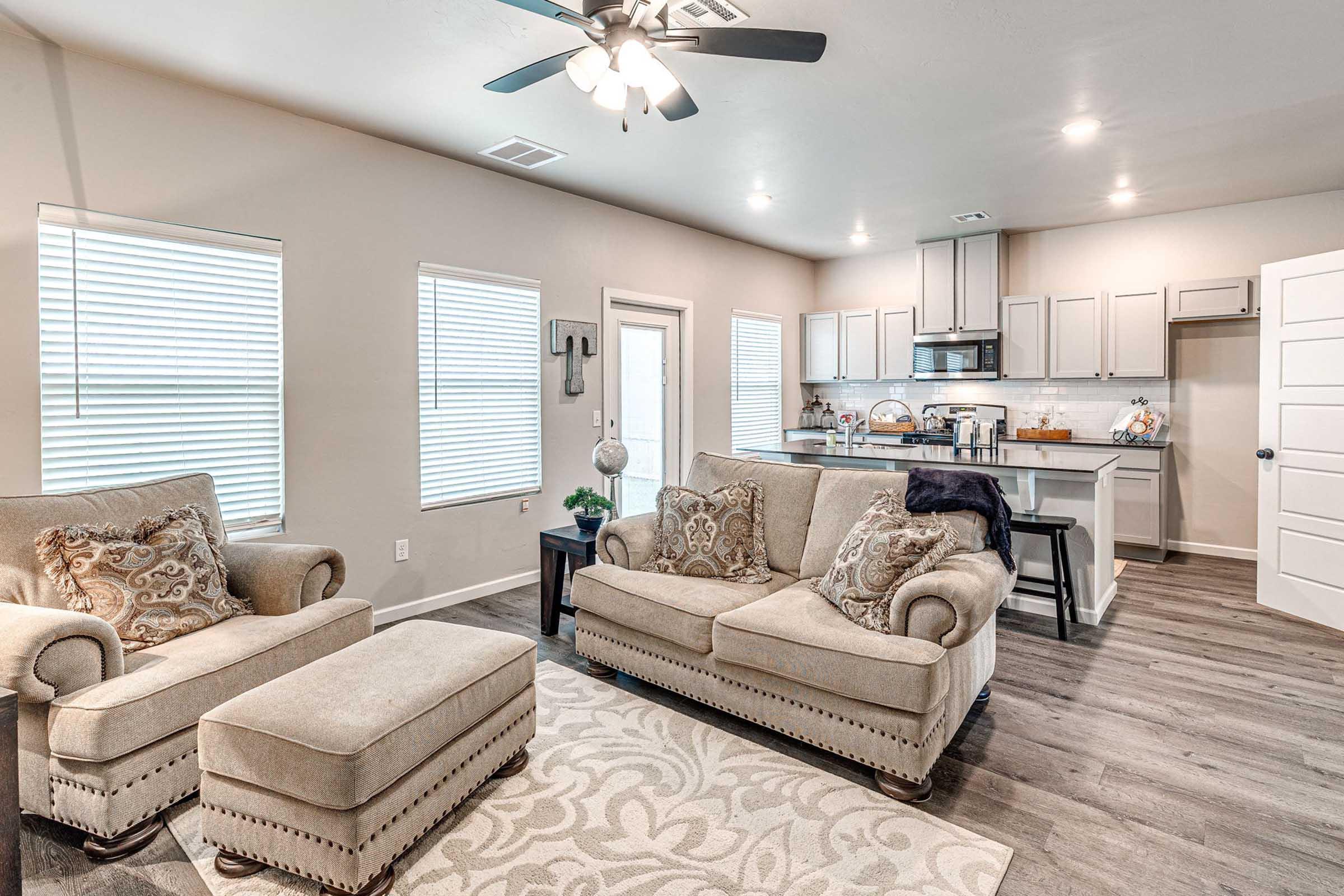 A cozy living room featuring two beige chairs with decorative pillows and an ottoman. Light-colored walls and wooden flooring create a warm ambiance. A kitchen area is visible in the background, equipped with modern appliances and a dining space. Natural light streams in through several windows.