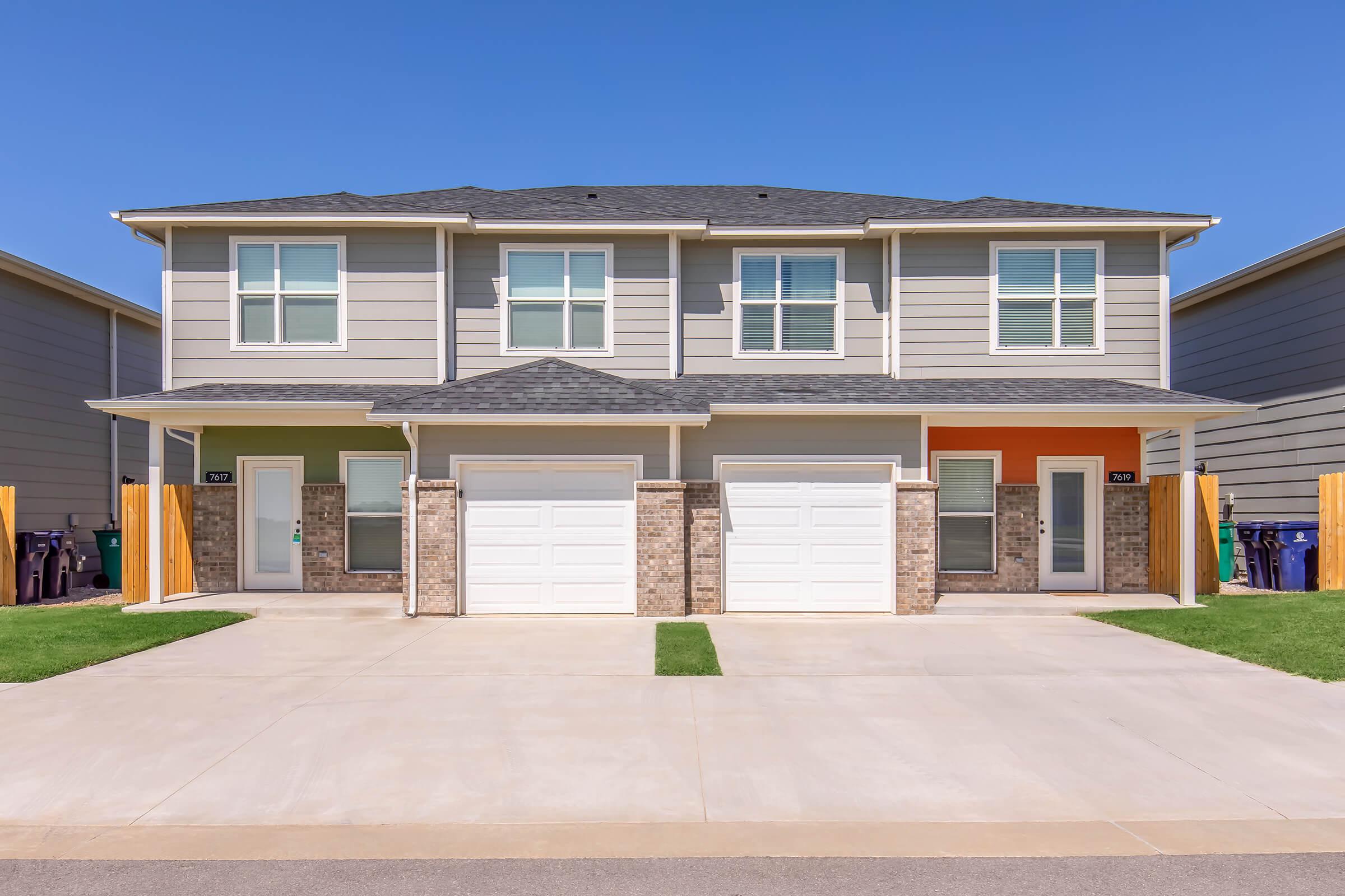 Two symmetrical townhome units with light gray siding, featuring brick accents and two-car garages. Each unit has a front door—one painted orange, the other in a neutral tone. The driveways are paved, with green grass lining the front. Clear blue sky is visible in the background.