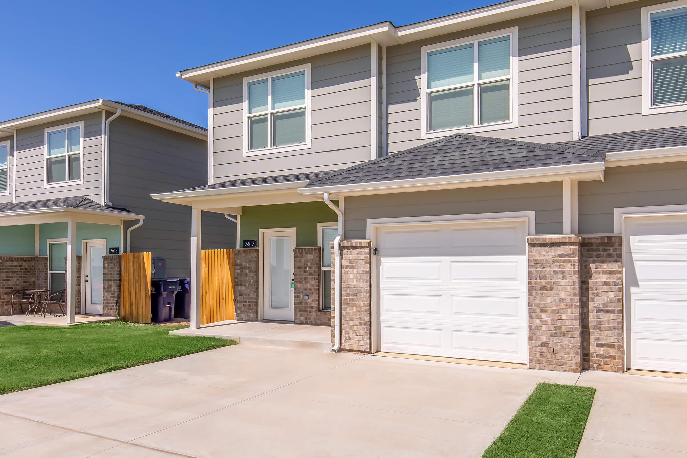 A modern two-story townhouse with a garage, featuring a gray exterior and a neatly maintained front yard. The driveway is paved, and there are adjacent homes visible. Bright blue sky overhead enhances the inviting appearance of the property.