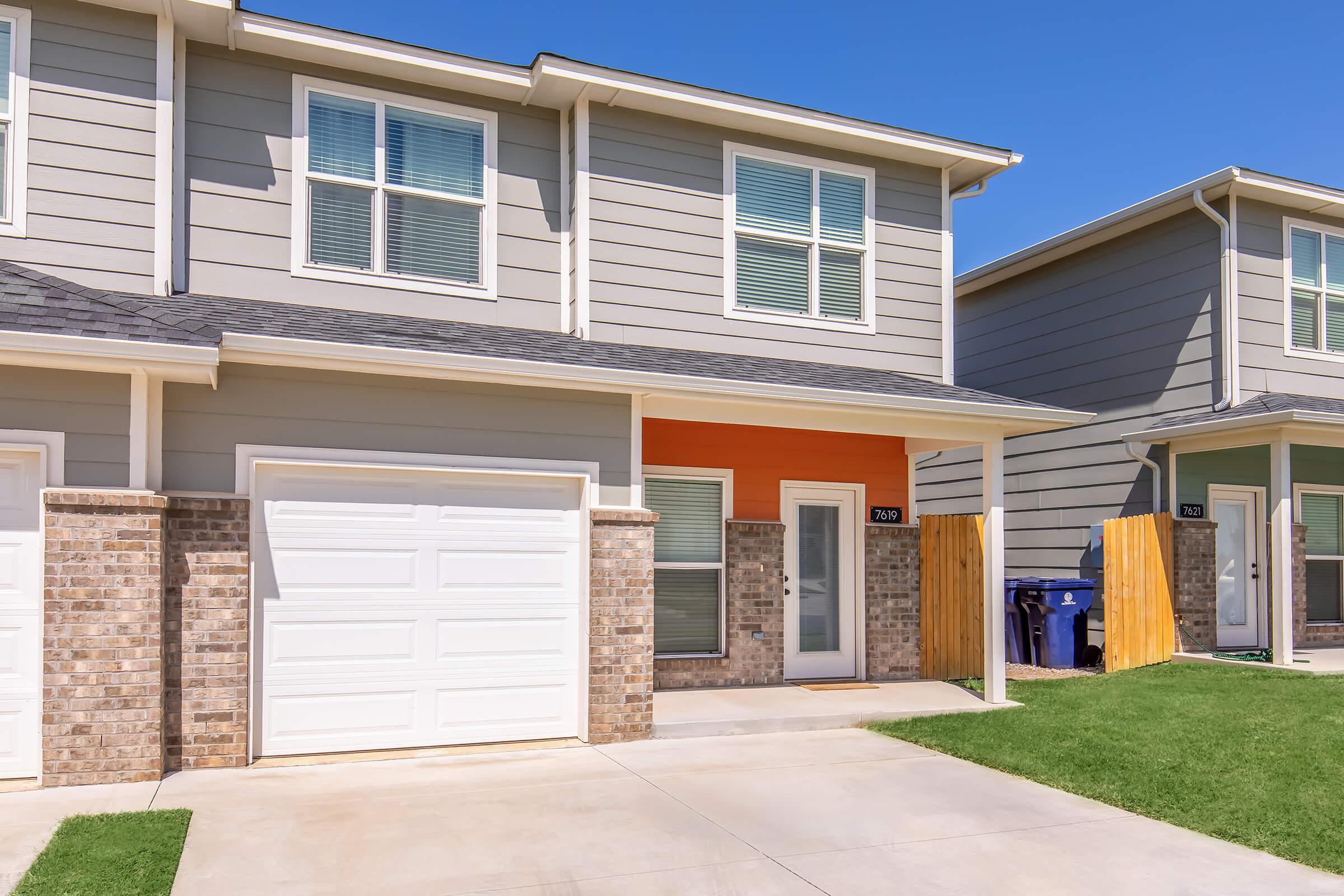 A modern two-story townhouse with a light gray exterior and a bright orange accent wall. The front features a garage door, a small porch area with a glass door, and neatly manicured grass. Adjacent to the property, a wooden fence and a blue trash bin are visible. Clear blue sky above.