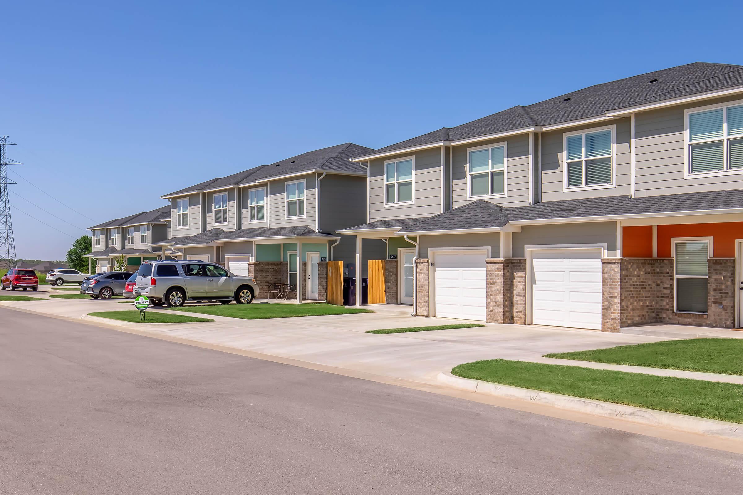 A suburban street view featuring a row of modern two-story townhouses with garages. The houses have a mix of grey siding and brick, with well-maintained lawns. Power lines are visible in the background, and parked vehicles can be seen along the street in clear weather.