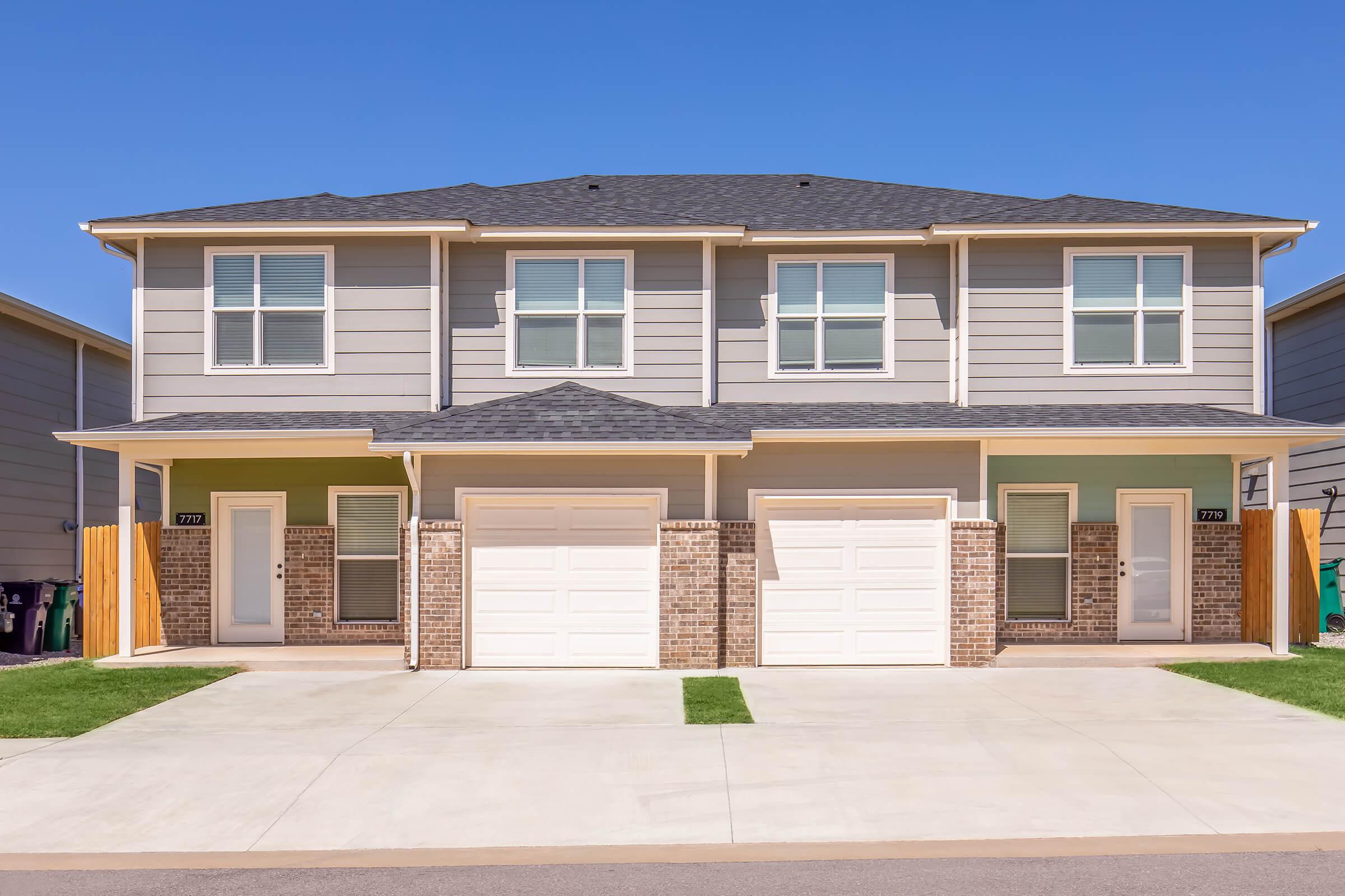 A two-story duplex with a beige exterior and brick accents, featuring two garages. Each unit has three windows on the top floor and a front door with a small covered entryway. A paved driveway leads to the garages, with a green lawn on either side and a clear blue sky in the background.