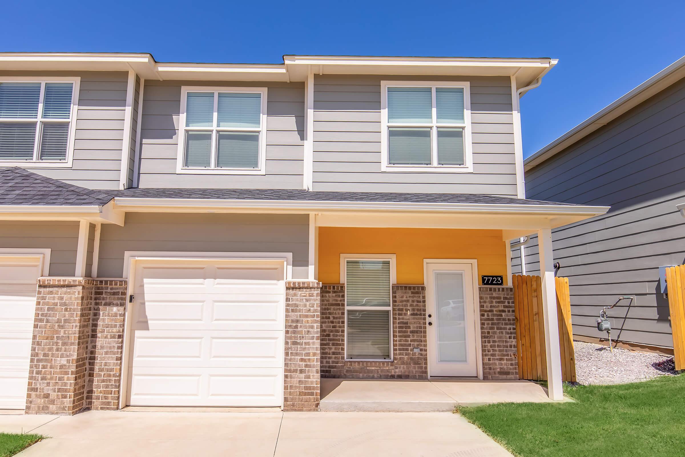 A modern two-story townhouse with gray siding and a vibrant orange accent wall. The front features a garage door on the left and a welcoming entrance with a white door. A small patch of lawn is visible, and there are wooden fences on either side, under a clear blue sky.