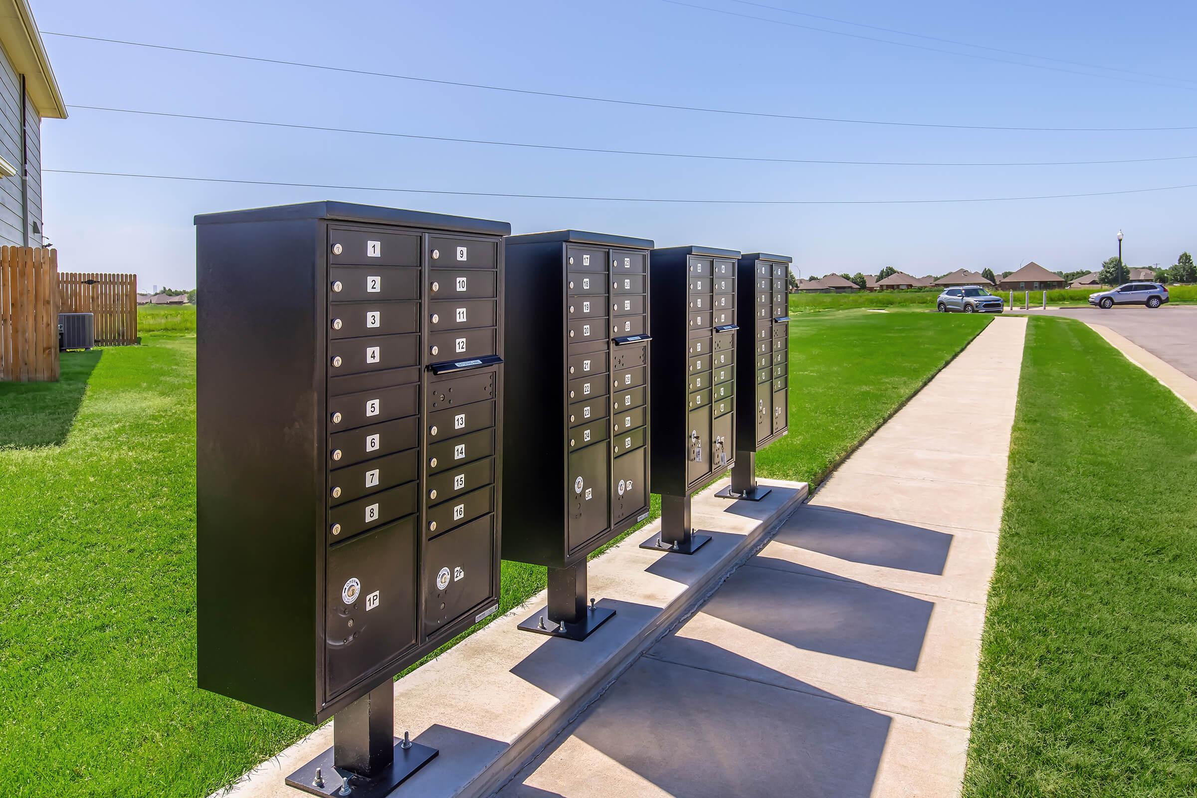 A row of modern black mailboxes situated on a paved pathway, surrounded by a grassy area under a clear blue sky. The mailboxes are organized in four units, each featuring multiple individual compartments for letters and packages.