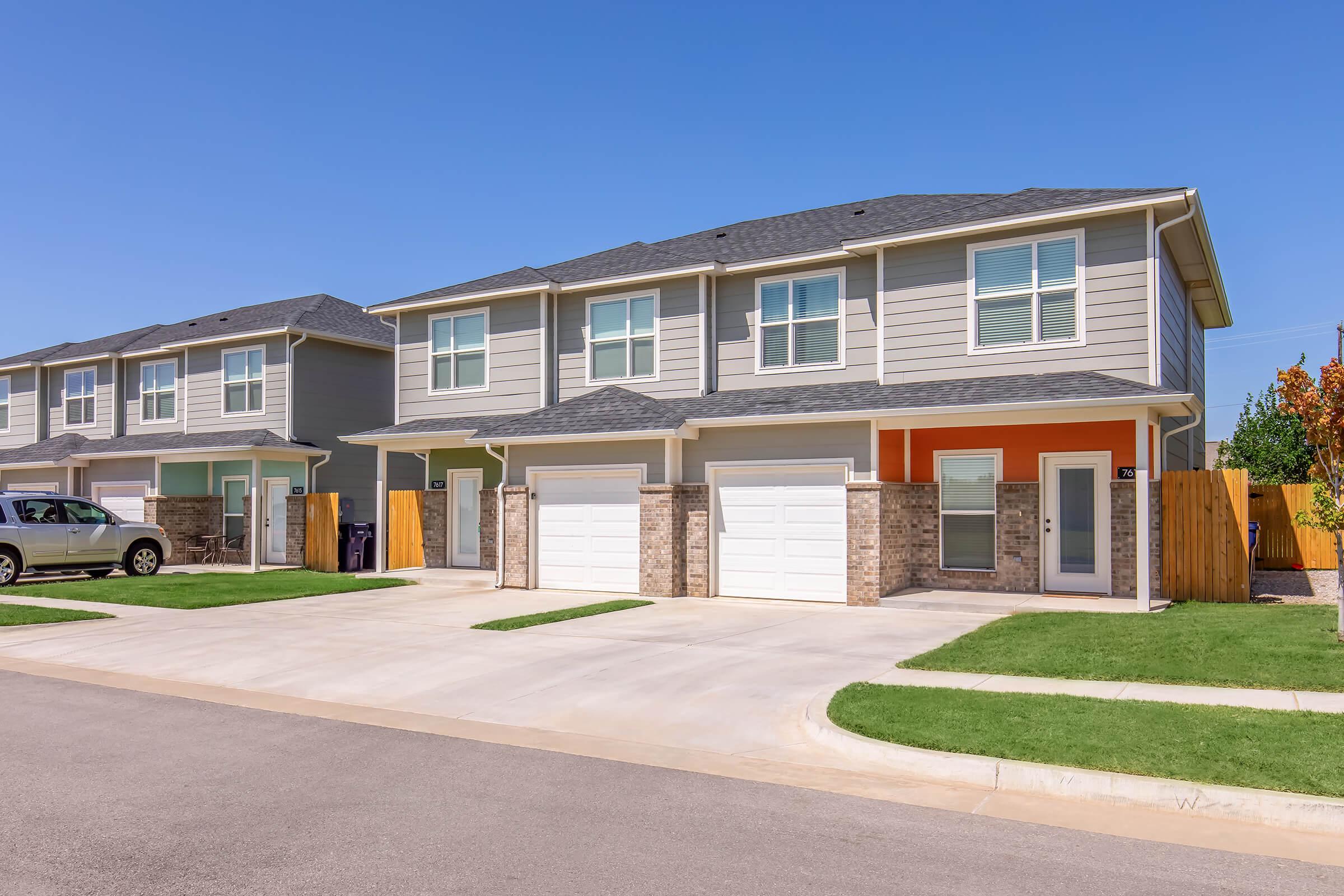 A modern duplex featuring two separate units, each with a two-car garage and a welcoming front porch. The exterior is painted in light gray with orange accents, surrounded by a well-maintained lawn and a paved driveway. Clear blue skies complete the inviting residential scene.