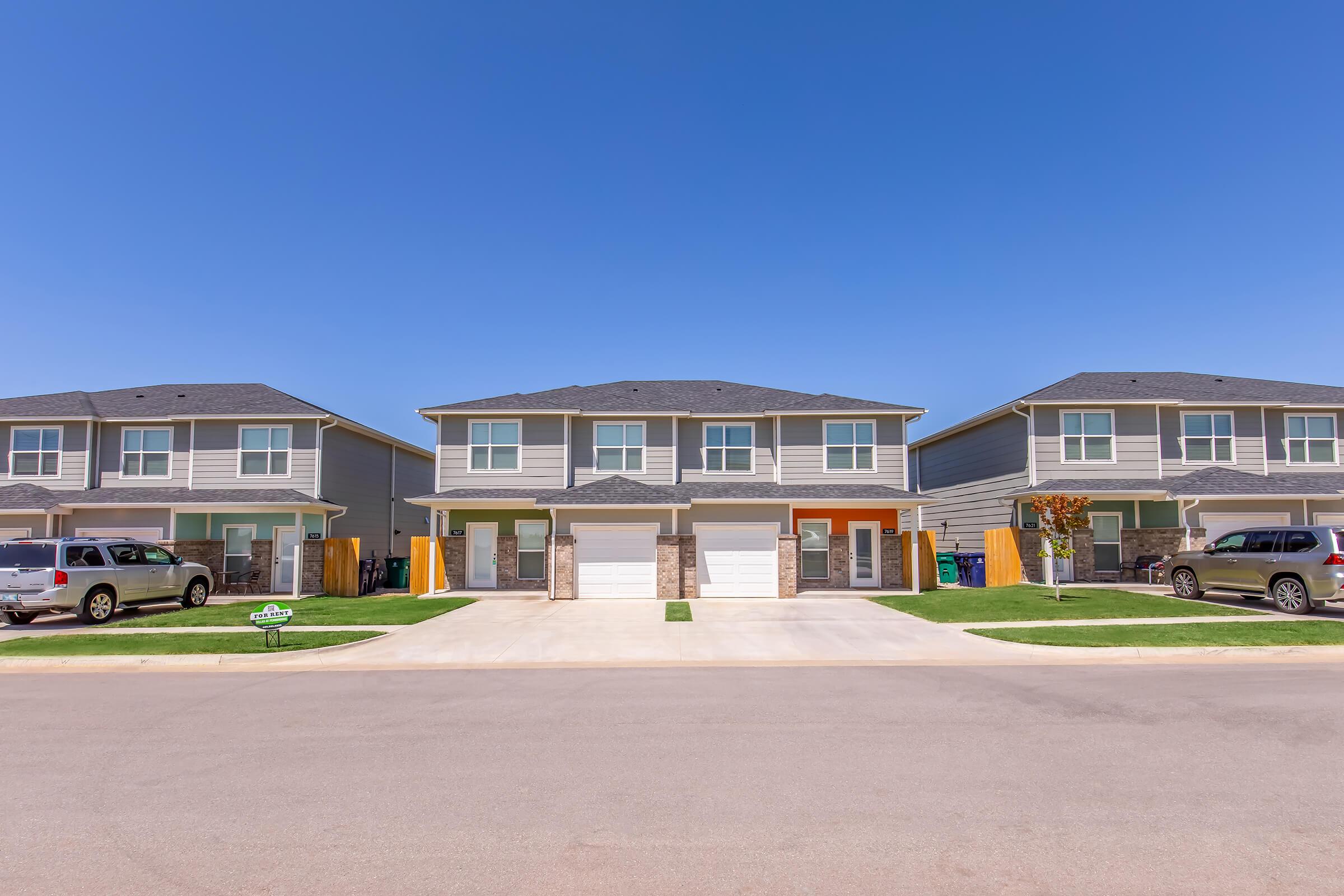 A row of modern duplex homes with gray siding and attached garages, set against a clear blue sky. Green lawns flank the paved driveway, and parked vehicles are visible on the left side. The neighborhood appears well-maintained and inviting.