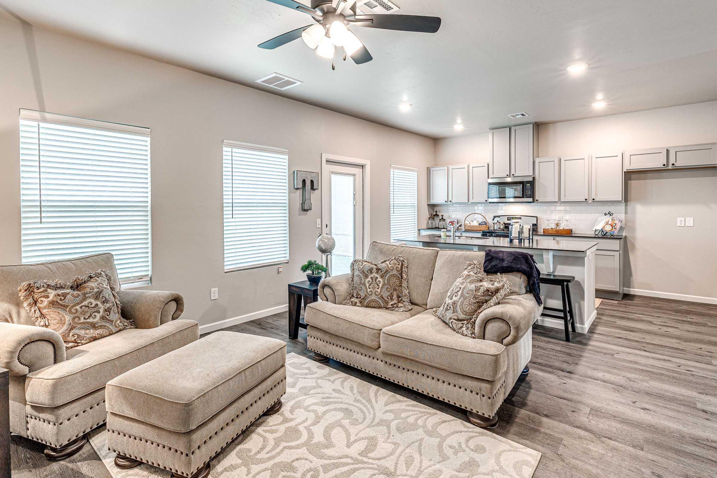 Cozy living room featuring two beige armchairs and a matching ottoman, adorned with decorative pillows. A light-colored area rug lies on the wooden floor. In the background, a modern kitchen with grey cabinets and stainless-steel appliances is visible, complemented by natural light from several windows.