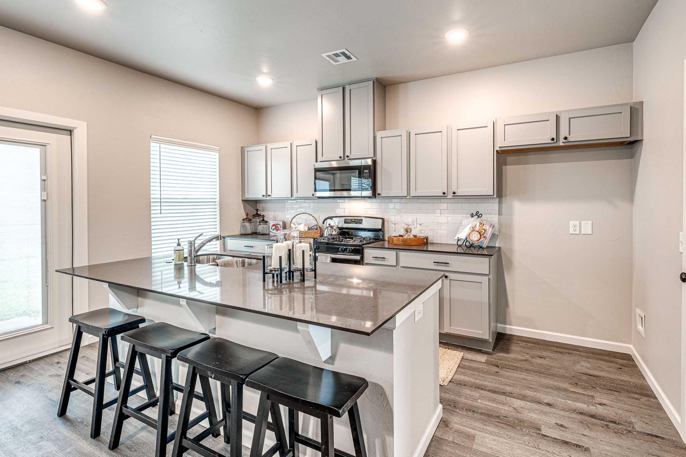 A modern kitchen featuring gray cabinetry, a sleek gray countertop, and a stainless steel stove. The kitchen island has black bar stools, and there is ample natural light coming through a window. The walls are painted a light color, and decorative items are placed on the countertop.