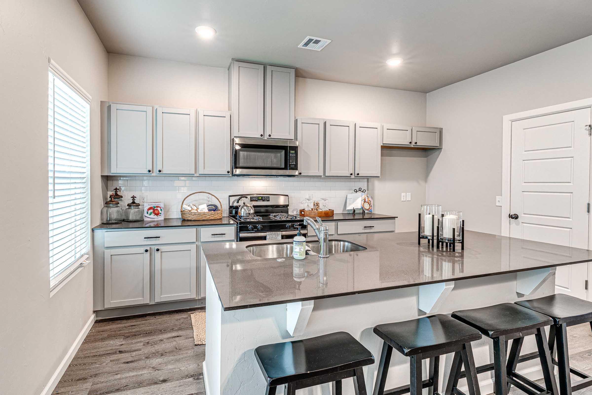 Modern kitchen featuring gray cabinets, stainless steel appliances, and a central island with bar stools. Natural light enters through a window, illuminating the space. The countertops are dark, contrasting with the light cabinetry, creating a contemporary aesthetic.