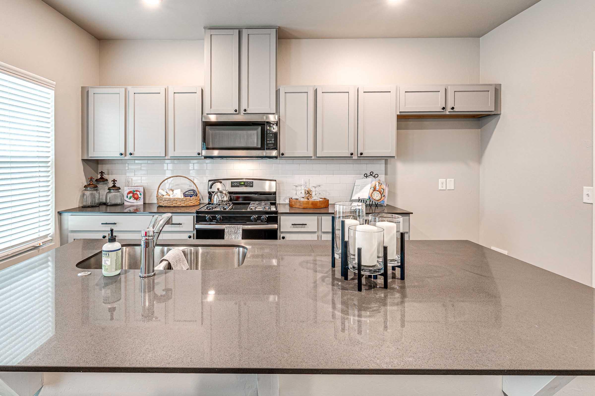 Modern kitchen featuring cabinets in light gray, a stainless steel stove and microwave, and a dark countertop with a sink. A decorative centerpiece with candles is placed on the island, and there’s natural light coming through the window. Minimalist decor enhances the inviting atmosphere.