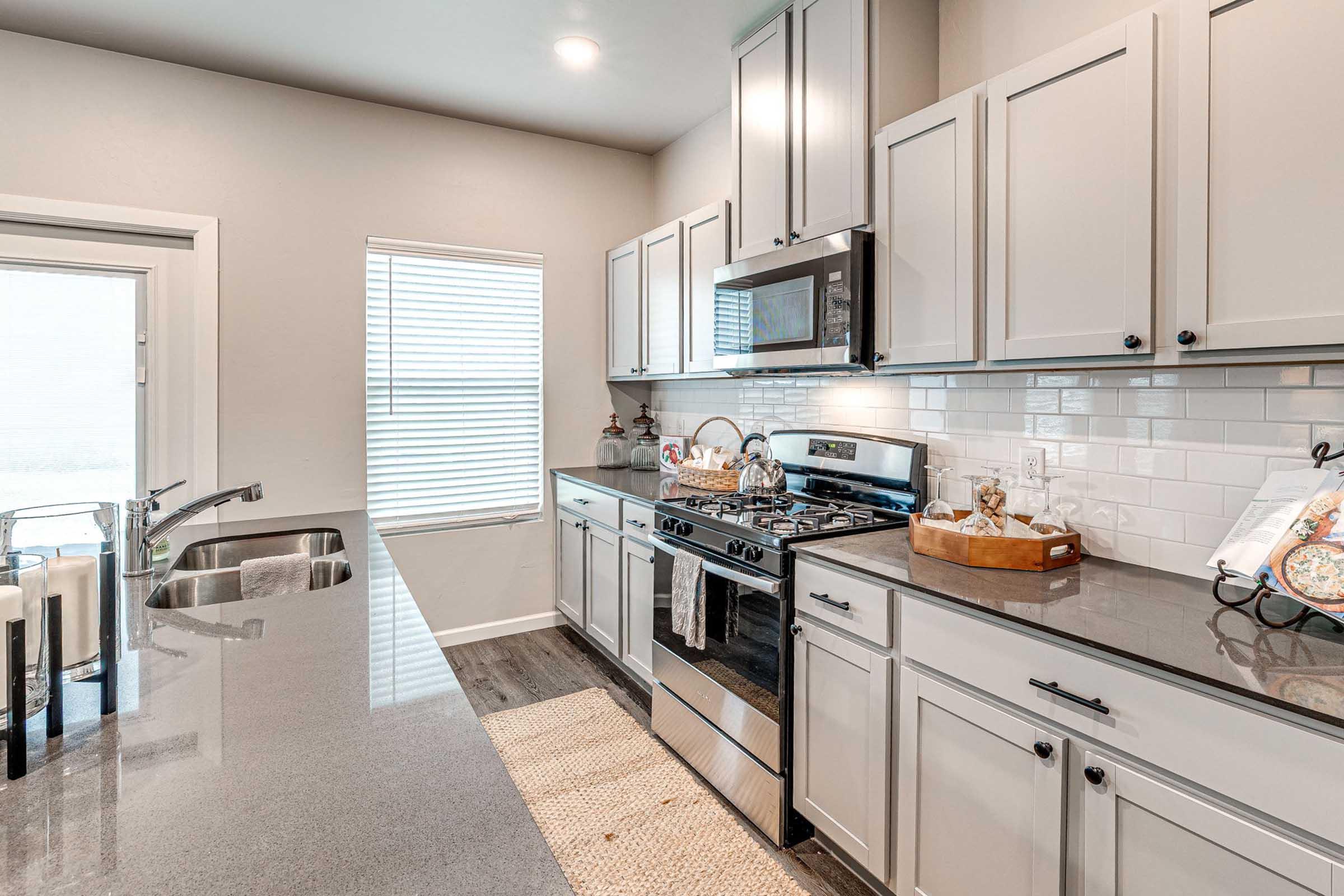 Modern kitchen featuring stainless steel appliances, a gas stove, and sleek gray cabinetry. The countertop is dark granite, with a matching backsplash. Natural light streams in through a large window next to the sink, and decorative items are arranged on the counter and shelves, creating a warm and inviting atmosphere.