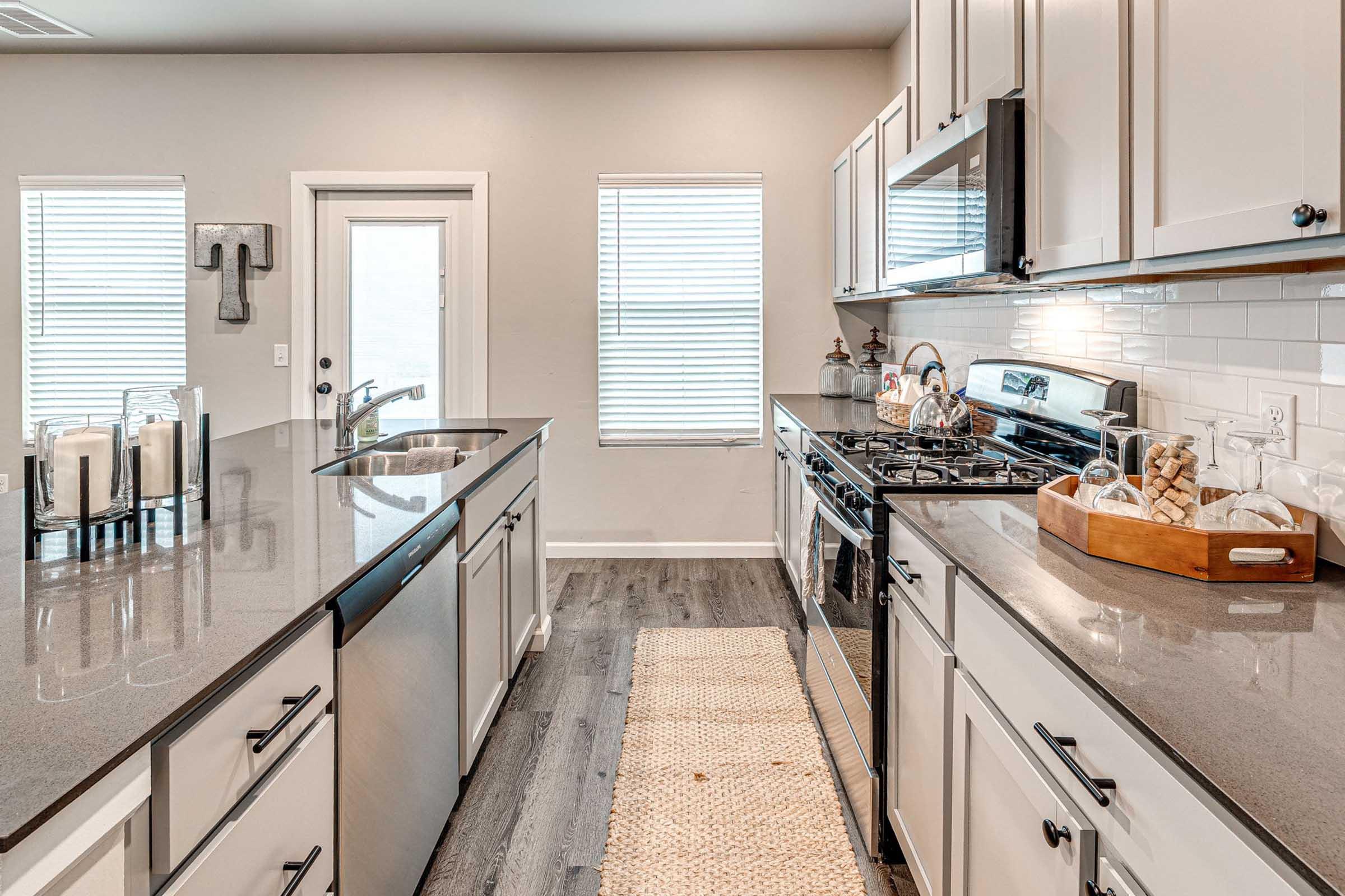 A modern kitchen featuring white cabinets, granite countertops, and stainless steel appliances. The space includes a large sink with a faucet, a stove, and a decorative wooden tray with kitchen items. There are two windows providing natural light, and a woven rug on the floor adds a cozy touch.