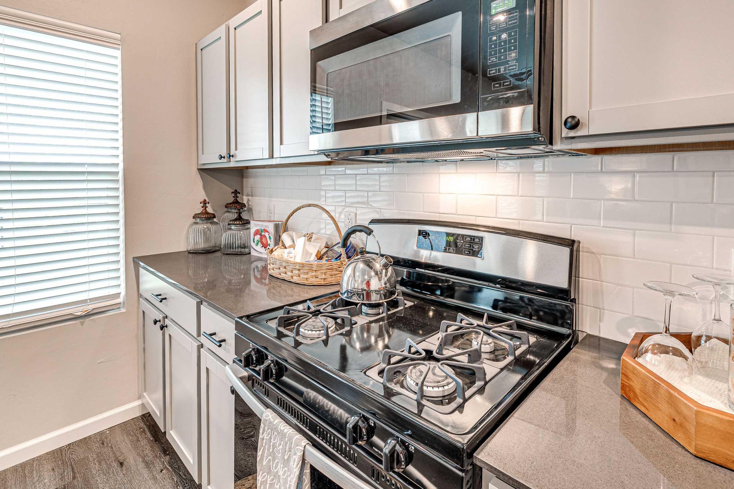 A modern kitchen featuring stainless steel appliances, including a microwave above a gas stove. The countertop is dark and sleek, with a basket containing utensils and a kettle on the stove. There's a window with blinds, allowing natural light to enter, and decorative elements like a lantern and a wooden box.