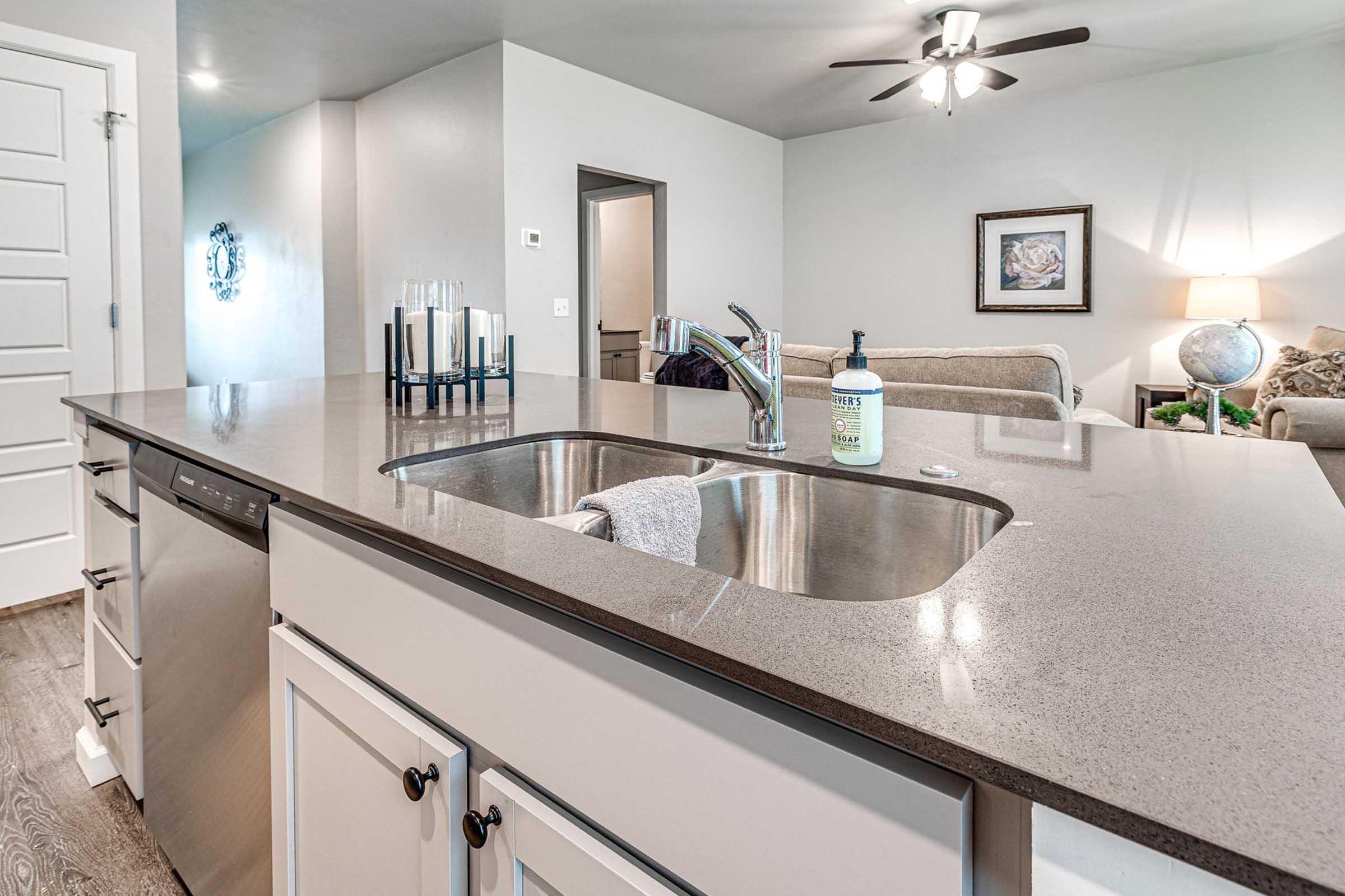 A modern kitchen featuring a granite countertop with a double sink and a stainless steel dishwasher. In the background, there's a cozy living area with a sofa, a lamp, and decorative items. The walls are painted a neutral color, and a ceiling fan is visible above.