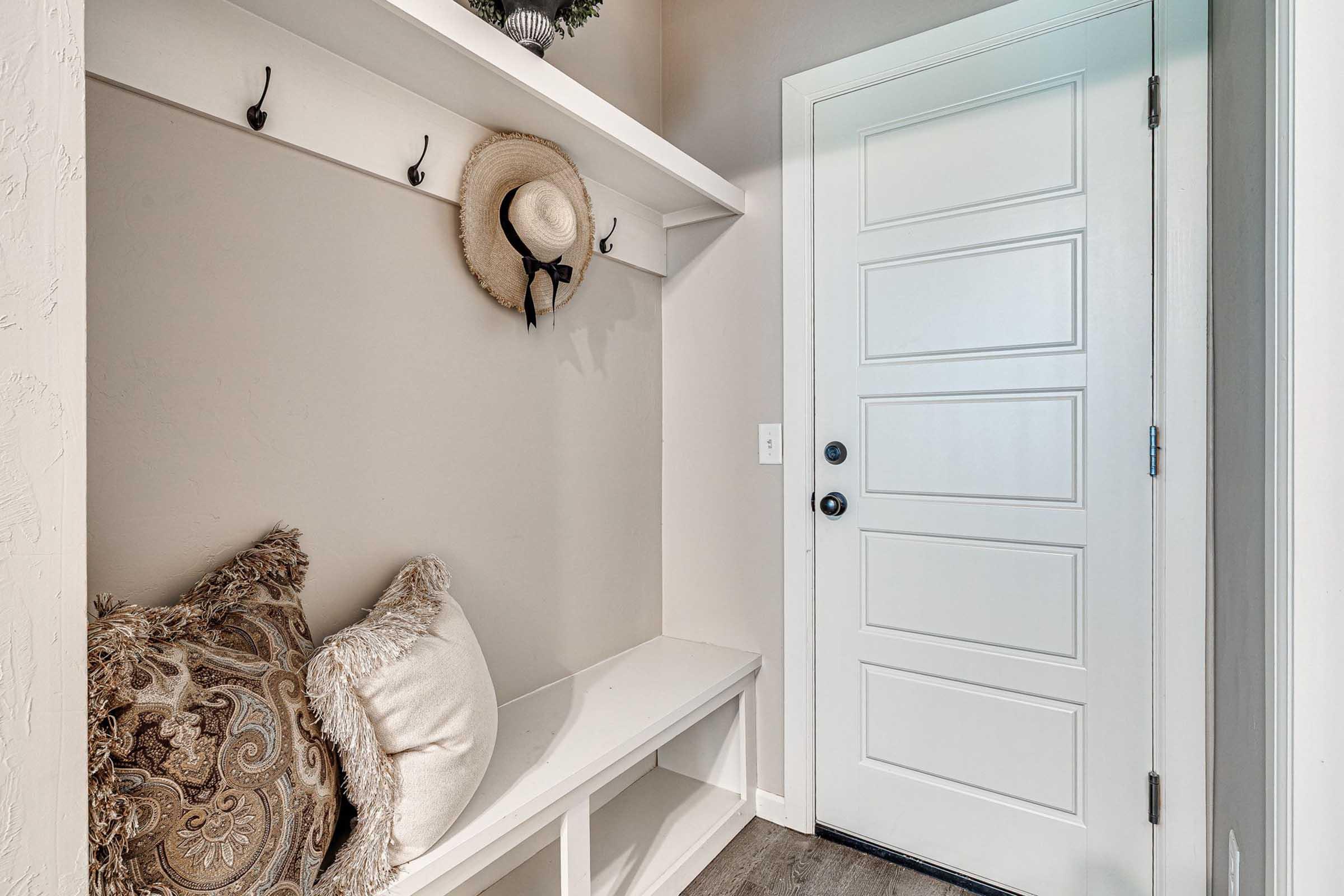 A cozy entryway featuring a white door, a bench with decorative pillows, and wall hooks. A straw hat hangs from one of the hooks, while a small decorative item sits on the shelf above. The walls are painted in a light color, creating a welcoming atmosphere.