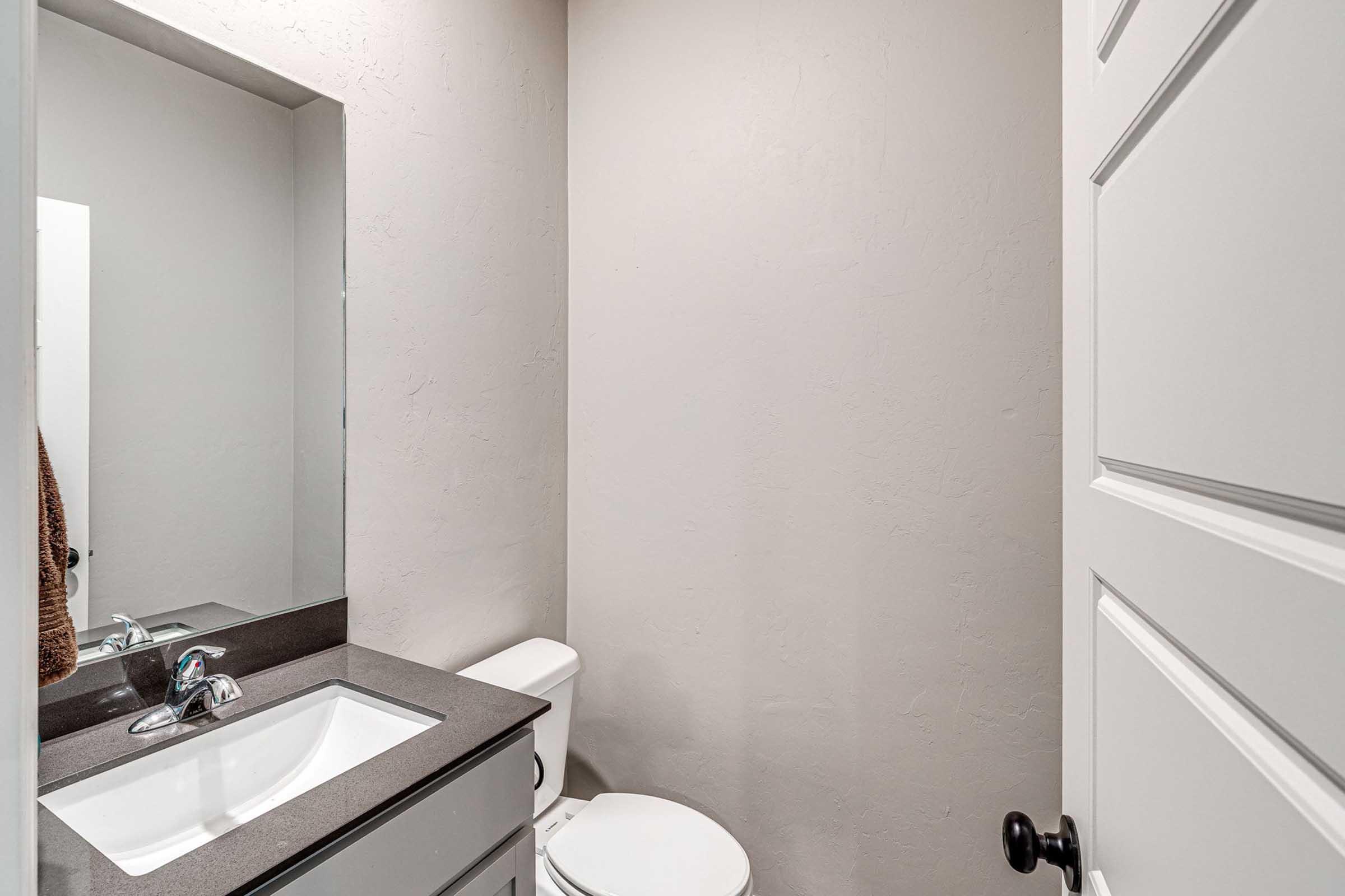 A small, modern bathroom featuring a white toilet, a sink with a sleek faucet, and a mirror mounted above the sink. The walls are painted in a neutral tone, and there is a black door handle for a wooden door. A towel is hanging near the sink. The overall design is clean and minimalistic.