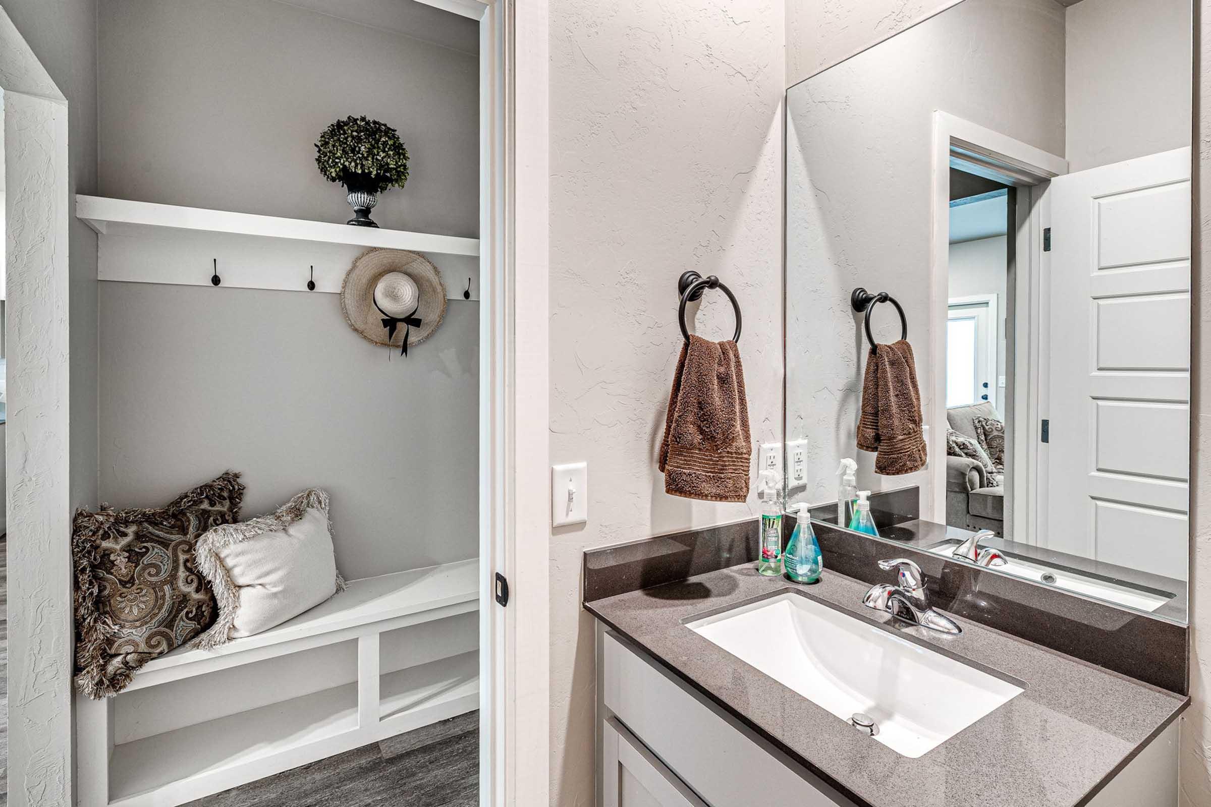 A modern bathroom featuring a sink with a polished countertop, a mirror, and stylish hand towels. To the side, there is a small entryway area with a bench, decorative hat, and a potted plant on a shelf, all set against light gray walls and a tidy, contemporary design.