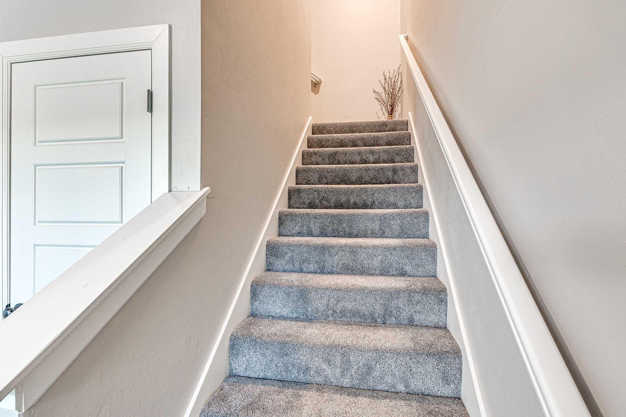 A softly lit staircase with gray carpet, leading up to a bright landing. White walls line the staircase, and a small decorative plant is placed on the landing. A closed white door is visible on the left side.