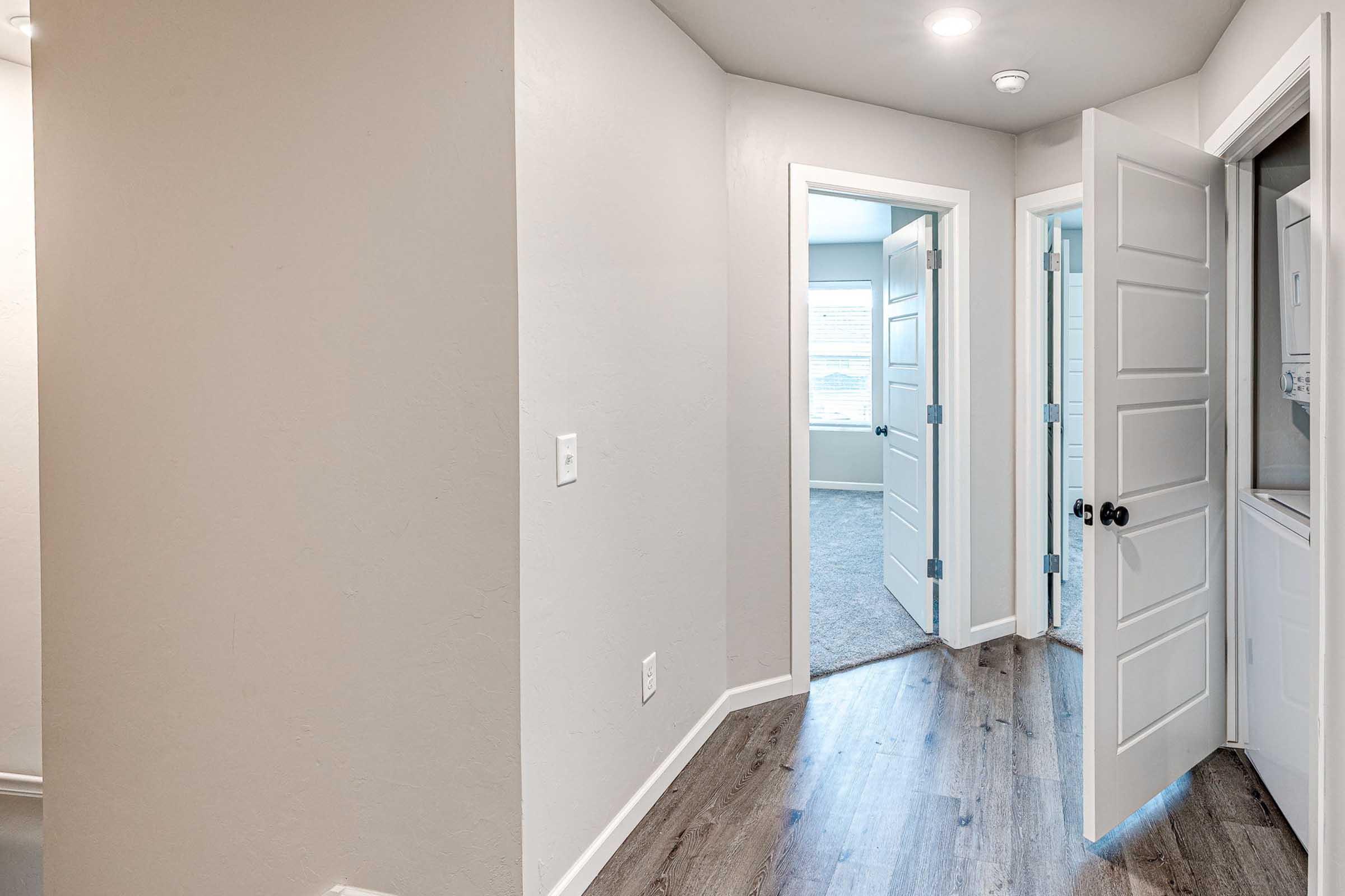 Light-colored hallway with smooth walls and a wooden floor. Multiple doors are visible, including one open door leading to another room and a door on the left side. The area is well-lit with overhead lighting, creating a spacious and inviting atmosphere.