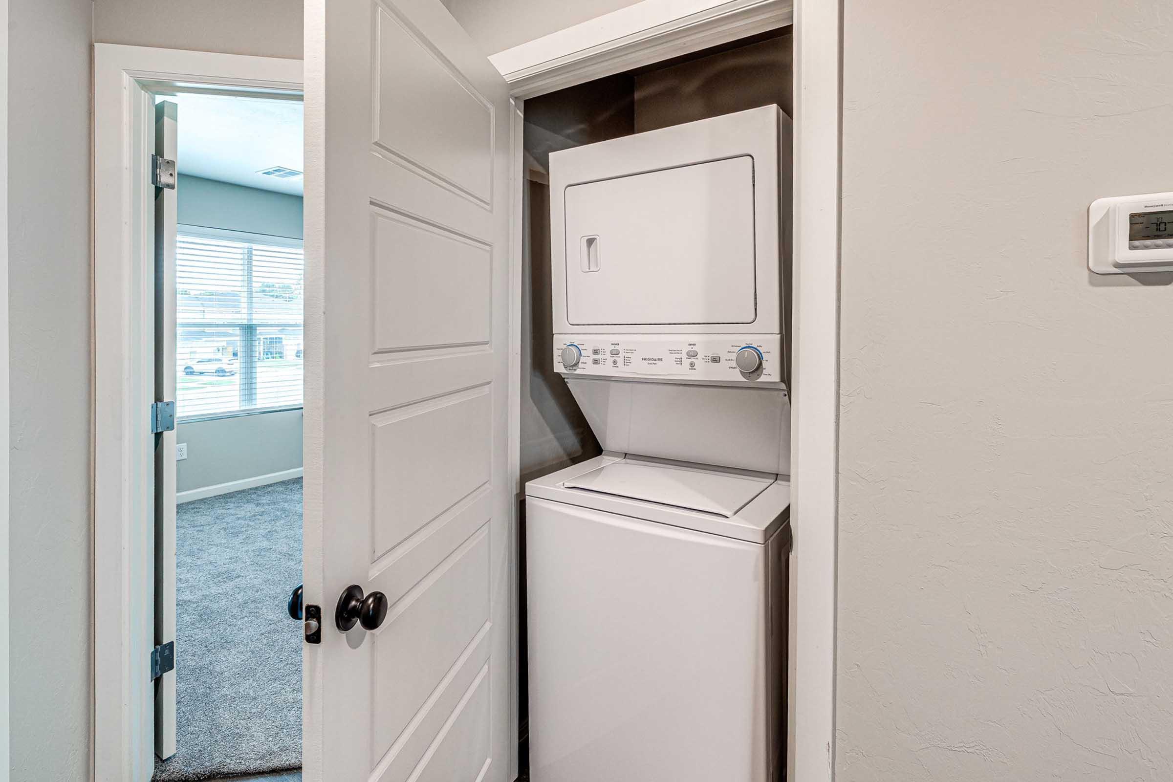 A compact laundry space featuring a stacked washer and dryer unit within a closet. The closet door is partially open, revealing the appliances. In the background, a bright room with large windows and carpet is visible, suggesting a modern and functional living space.