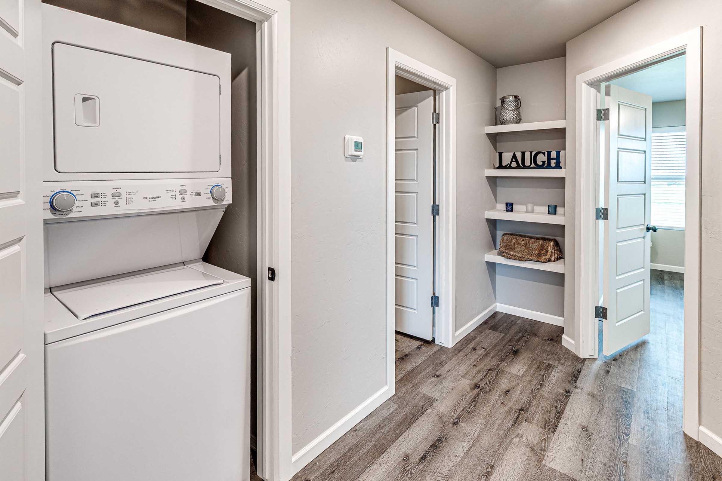 A modern laundry area with a stacked washer and dryer in an alcove. The hallway features two doors leading to separate rooms, light-colored walls, and wood-like flooring. A small shelf on the right has decorative items, including a sign that says “LAUGH.” Natural light coming from a window at the far end of the hall.