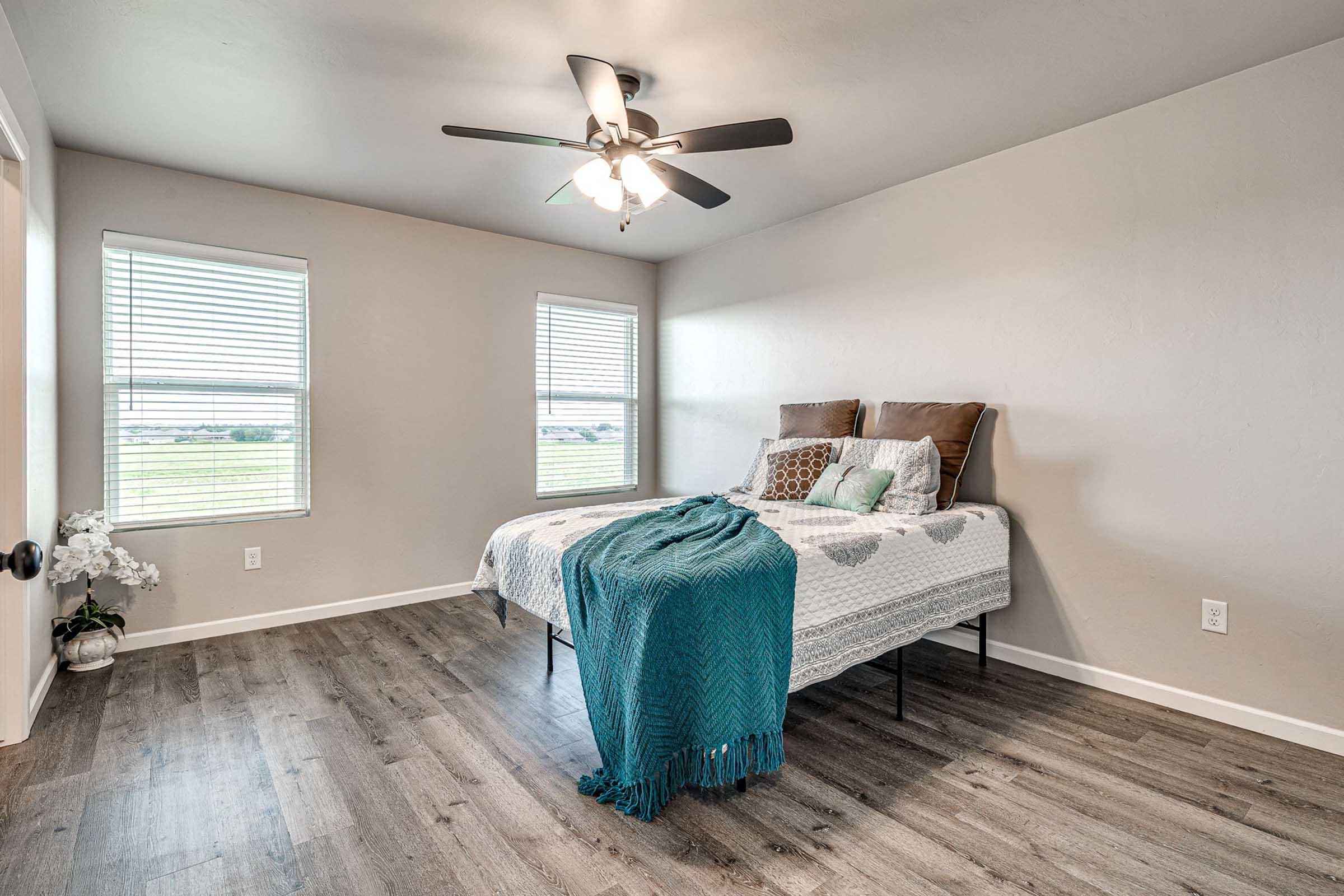 A bright bedroom featuring a bed with decorative pillows, a cozy blanket draped over it, and two windows allowing natural light. The floor is made of wood, and there is a potted plant beside the bed. A ceiling fan provides ventilation in the spacious, neutral-toned room.