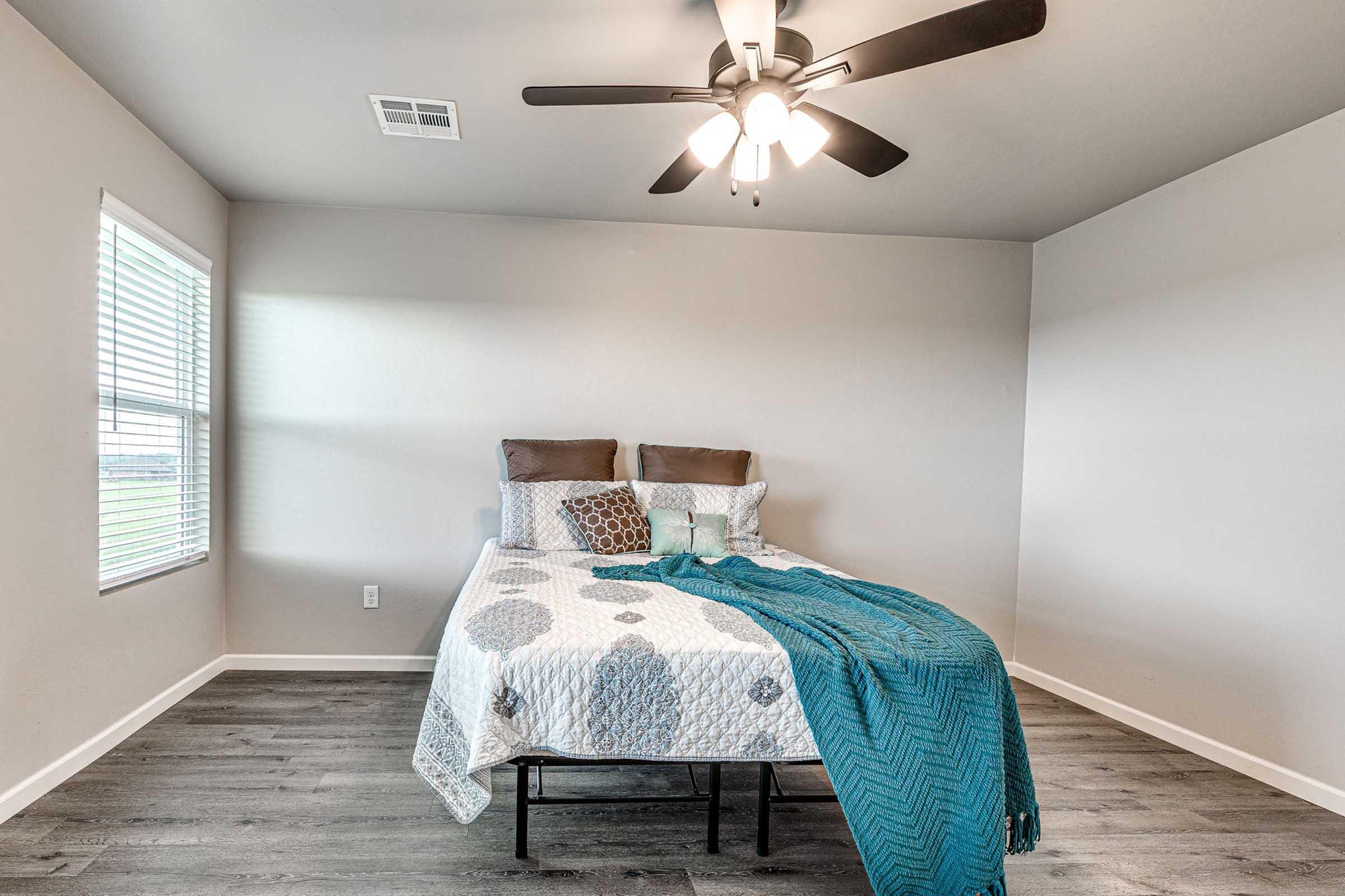 A well-lit bedroom featuring a bed with a patterned quilt and decorative pillows, a teal throw blanket draped across the foot, and a ceiling fan. There are two windows allowing natural light, with light-colored walls and wooden flooring completing the cozy atmosphere.