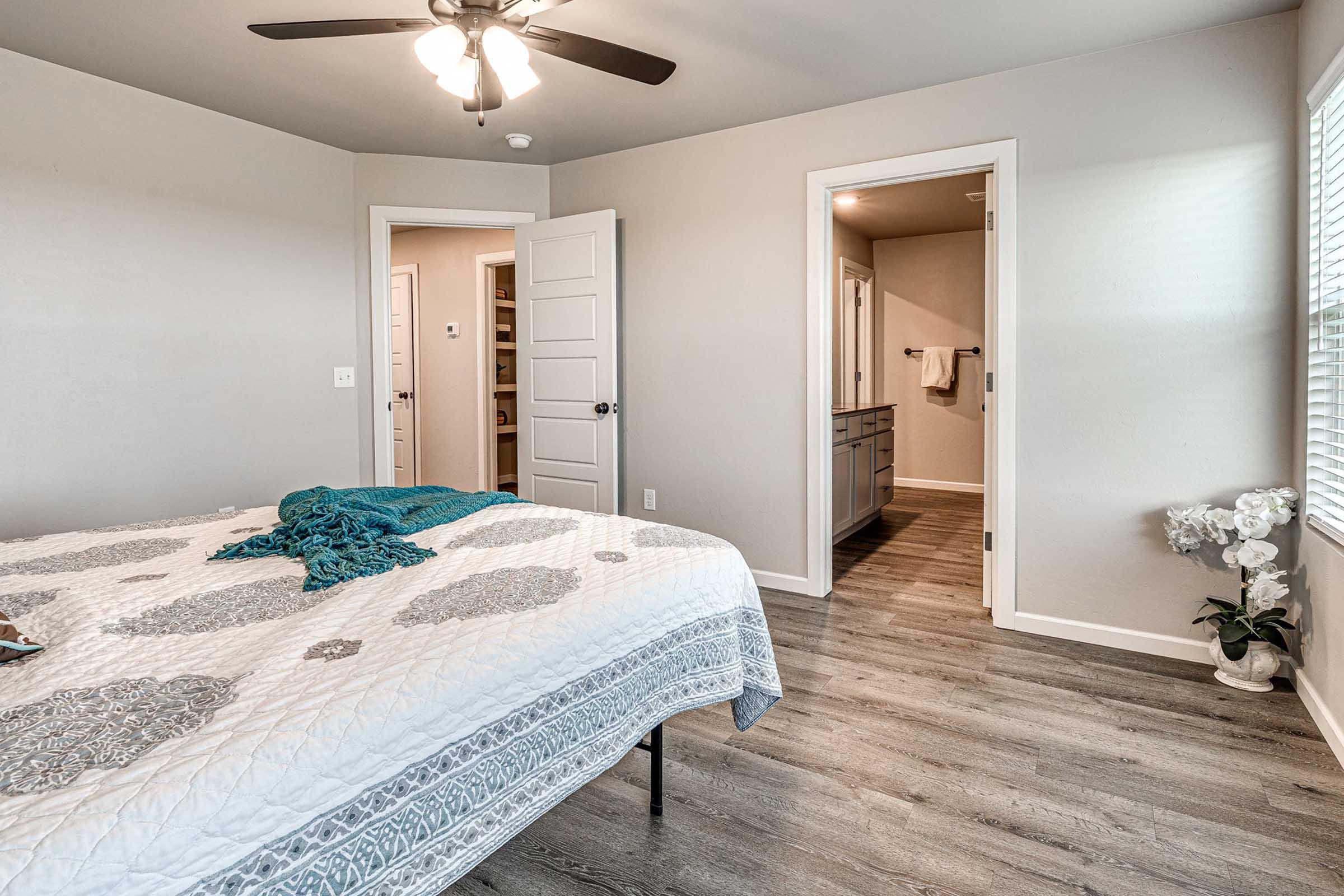 A cozy bedroom featuring a bed with a patterned quilt, a ceiling fan, and light-colored walls. A door leads to a bathroom, while another opens to a hallway with additional rooms. A potted plant sits next to the bed, contributing to the room's inviting atmosphere.