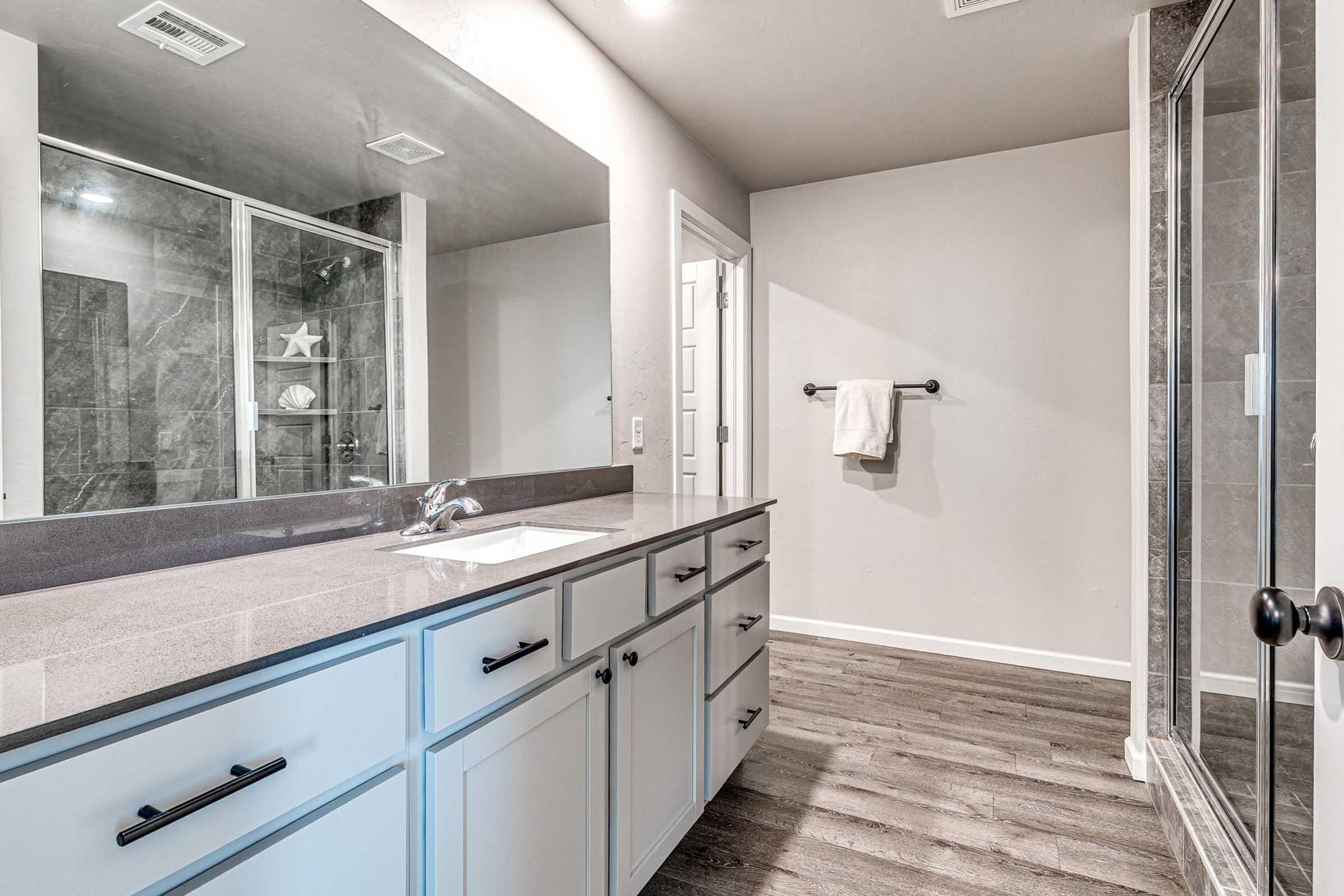 Modern bathroom featuring a clean design with a double vanity, sleek gray countertop, and large mirror. There is a glass-enclosed shower with gray tiles and a towel hanging on a rack. The flooring is a dark, wood-like material, adding warmth to the space. The walls are painted in a light, neutral color.