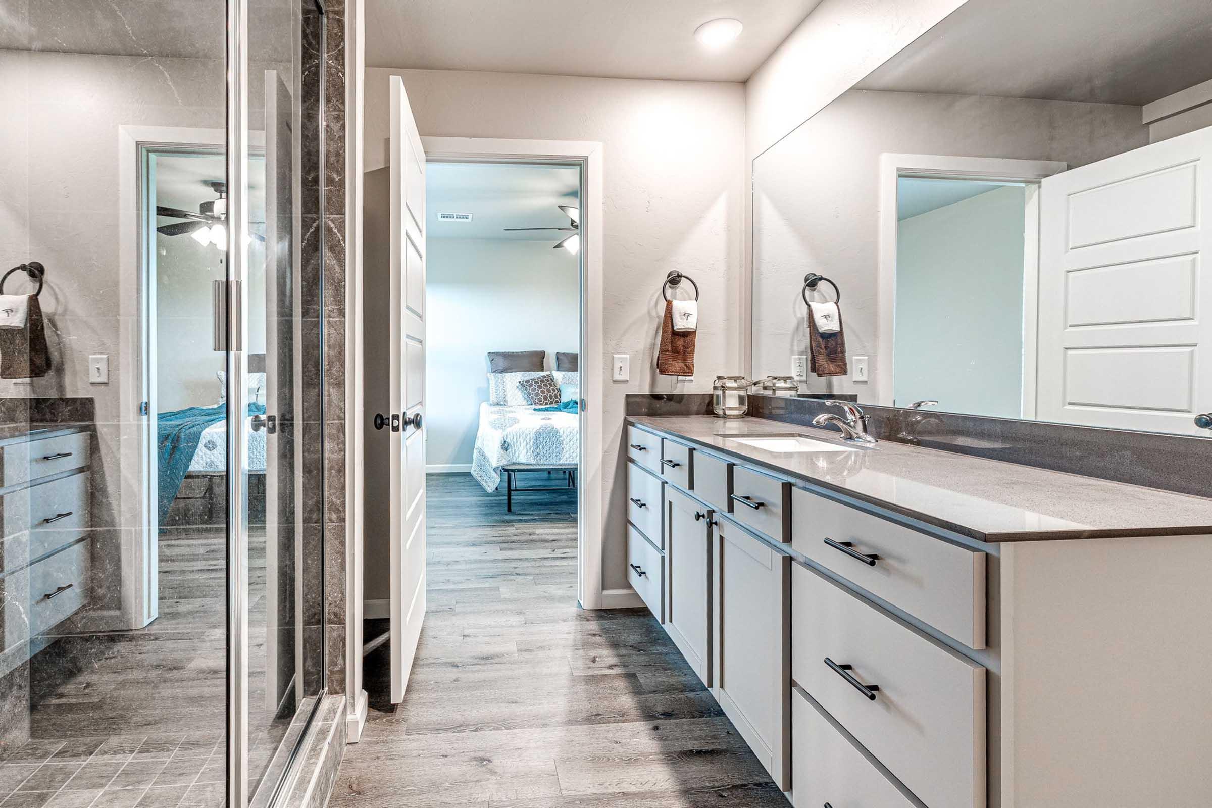 A modern bathroom featuring a double sink vanity with sleek cabinetry, a walk-in shower with glass doors, and light-colored walls. The space is well-lit with natural light visible through an adjacent room, which includes a bed and decorative bedding. The flooring is a warm-toned wood laminate.