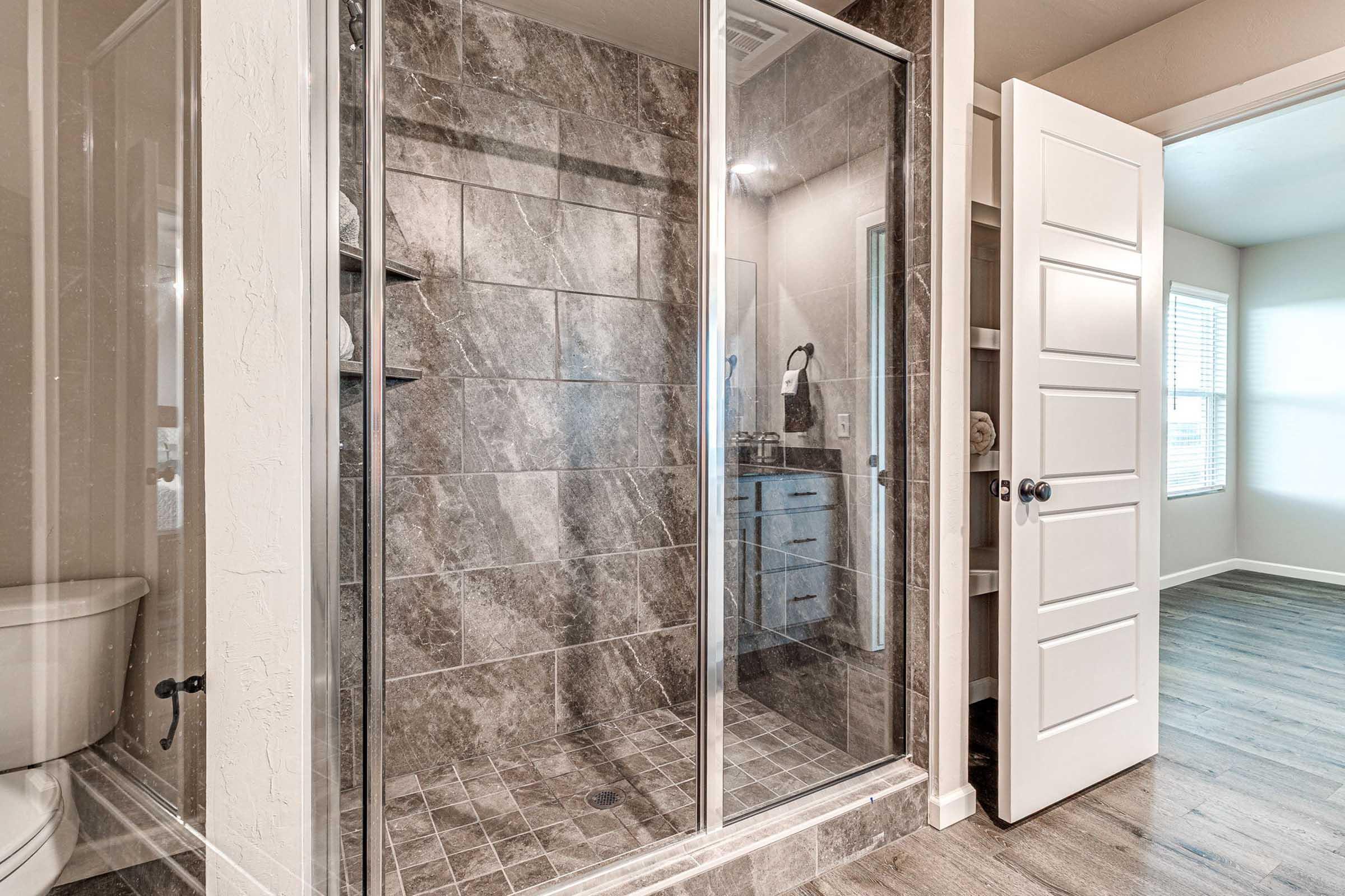 Modern bathroom featuring a glass-enclosed shower with tiled walls, a toilet, and a wooden floor. A white door leads to another room with natural light coming through a window. The shower area is clean and stylish, showcasing contemporary design elements.