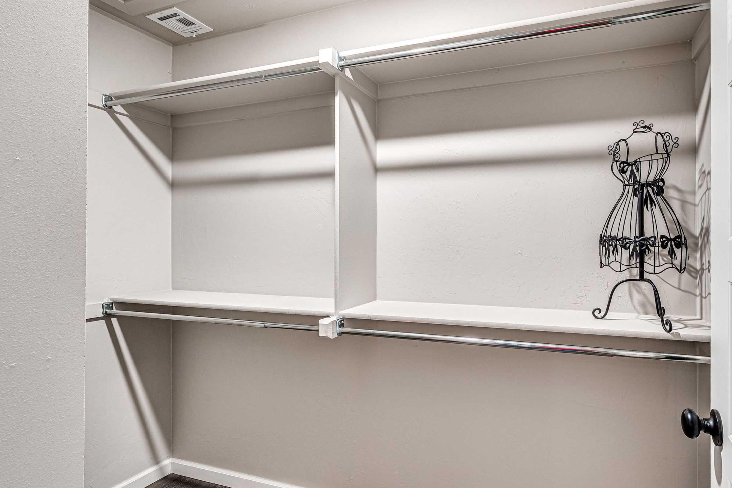 Empty closet with two metal shelves and a decorative wire dress form on the right side. The walls are light-colored, and the floor is dark wood. Natural light casts shadows, creating a clean and simple aesthetic.