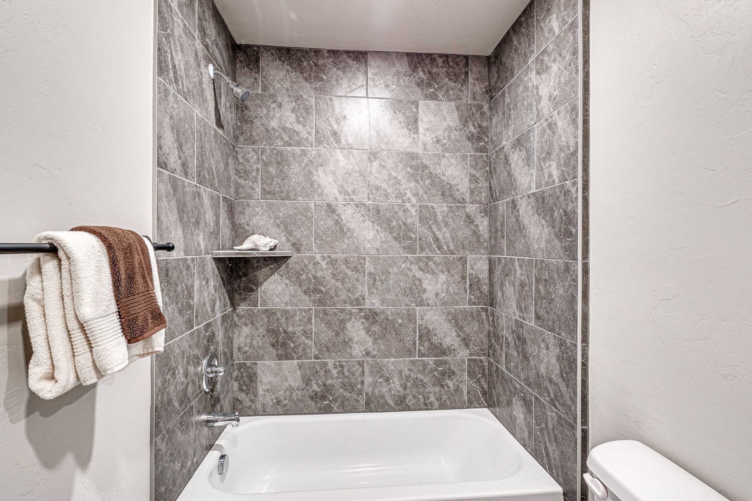 A modern bathroom featuring a bathtub with a gray tiled wall. The space includes a showerhead, a towel rack with a folded towel, and a small shelf with a washcloth. The overall design is sleek and contemporary, with neutral colors and clean lines.