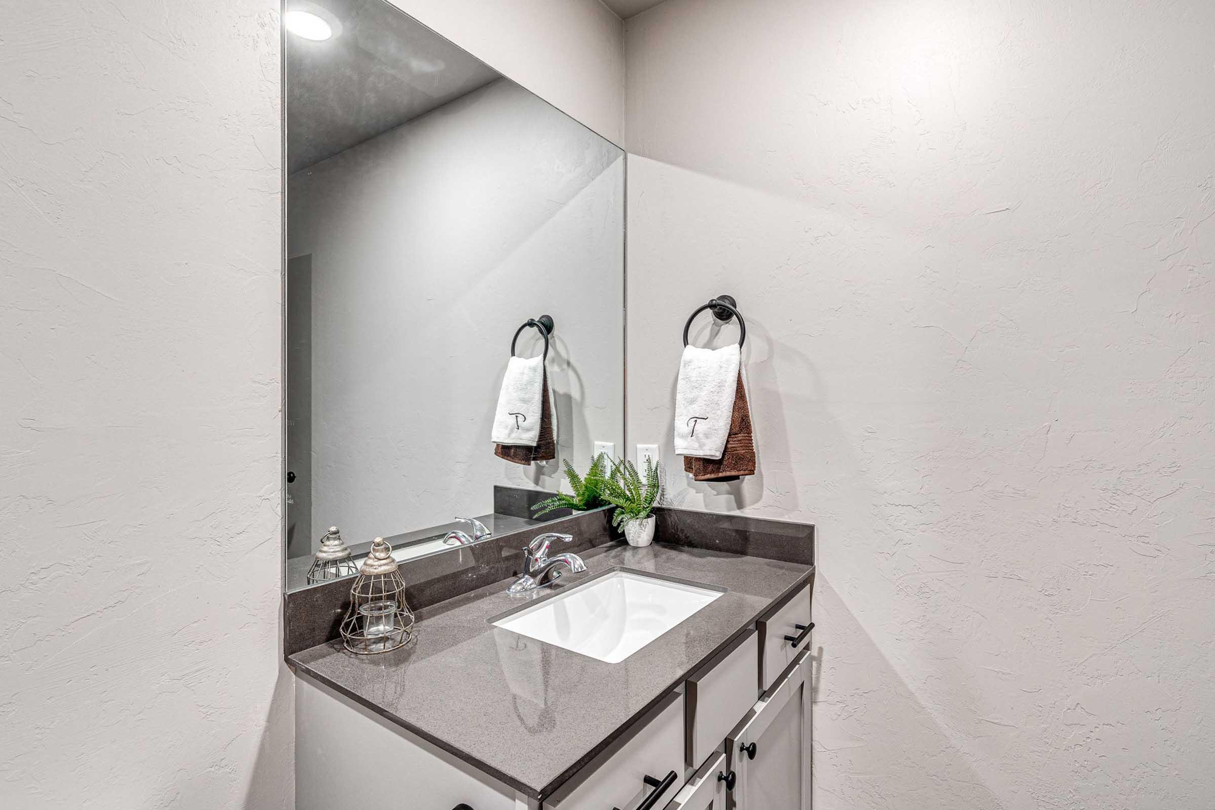 A modern bathroom featuring a sleek vanity with a sink, a large mirror above it, and decorative towels hanging on the wall. The countertop has subtle decor, including a small plant, adding a touch of greenery to the neutral-toned space. The overall design emphasizes cleanliness and simplicity.
