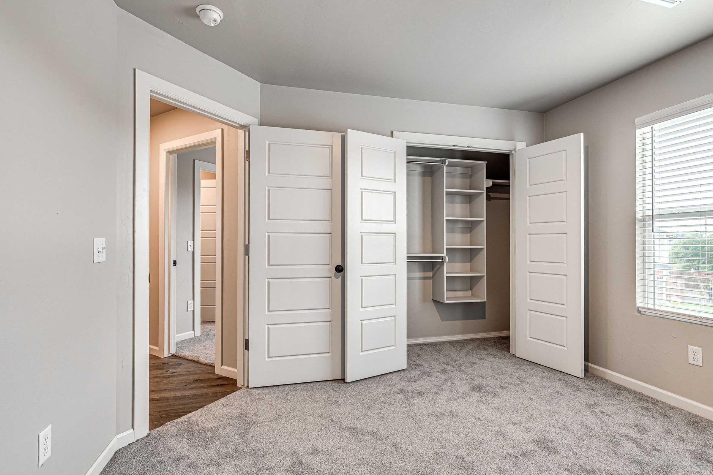 A well-lit room featuring two open double doors leading to a closet with shelving. The walls are painted in a soft gray, and the flooring is carpeted. Additional doors can be seen in the background, suggesting access to other areas of the home. Natural light filters through a window, enhancing the airy feel of the space.