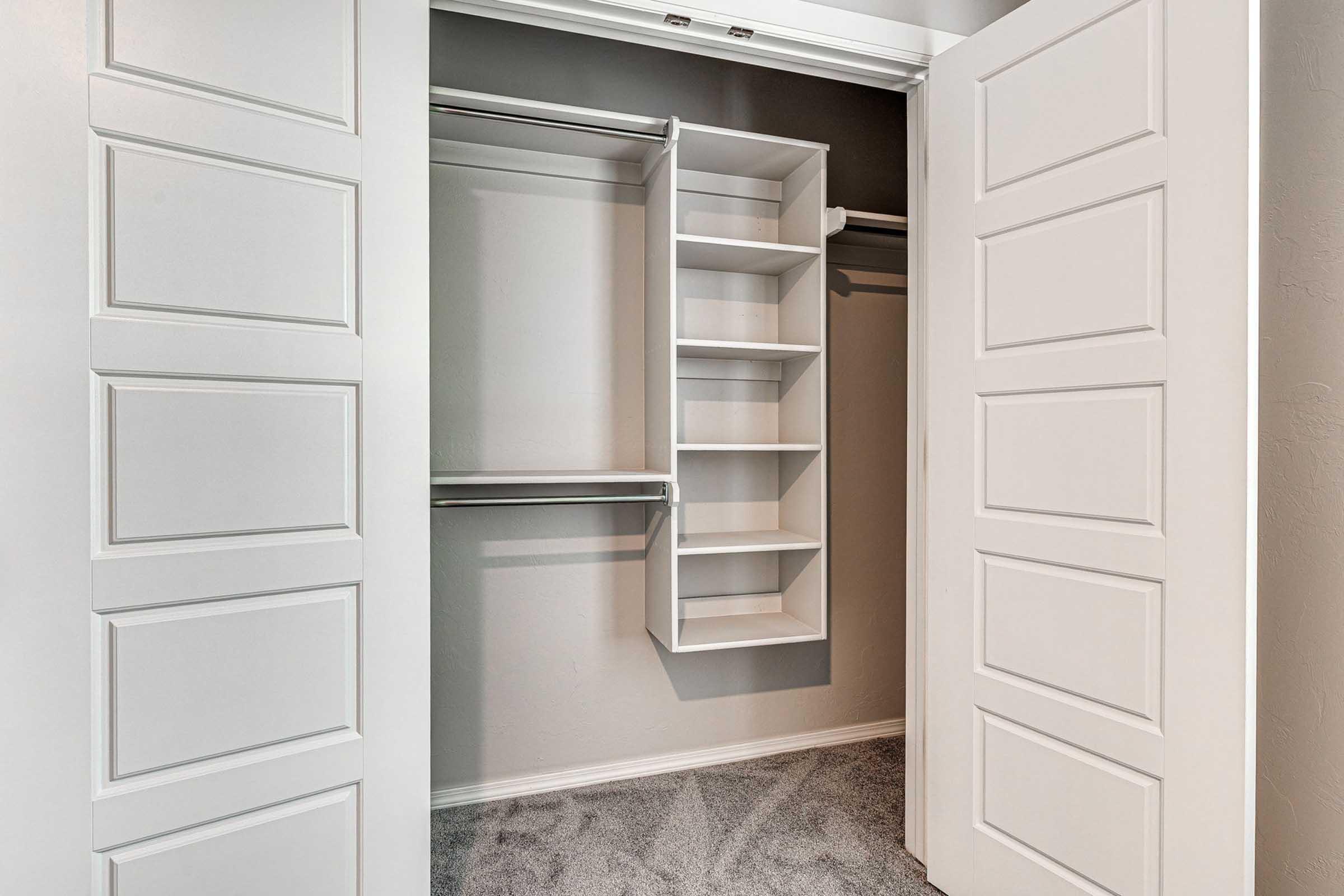 Empty closet with double doors open, revealing white shelving units and hanging space. The interior features light gray walls and carpet flooring, creating a clean and organized appearance.