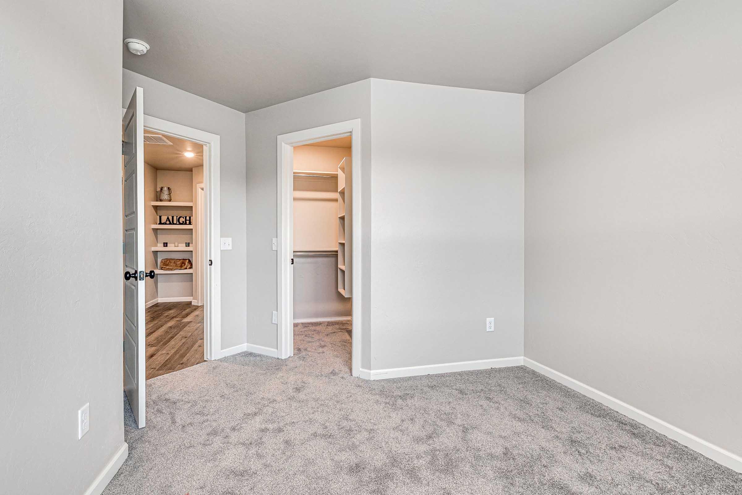 A light-colored room with carpet and a single open door leading to a closet space. The walls are painted in a neutral tone, and there is a second doorway leading to another area. The room appears spacious and well-lit, showcasing clean lines and minimal decor.