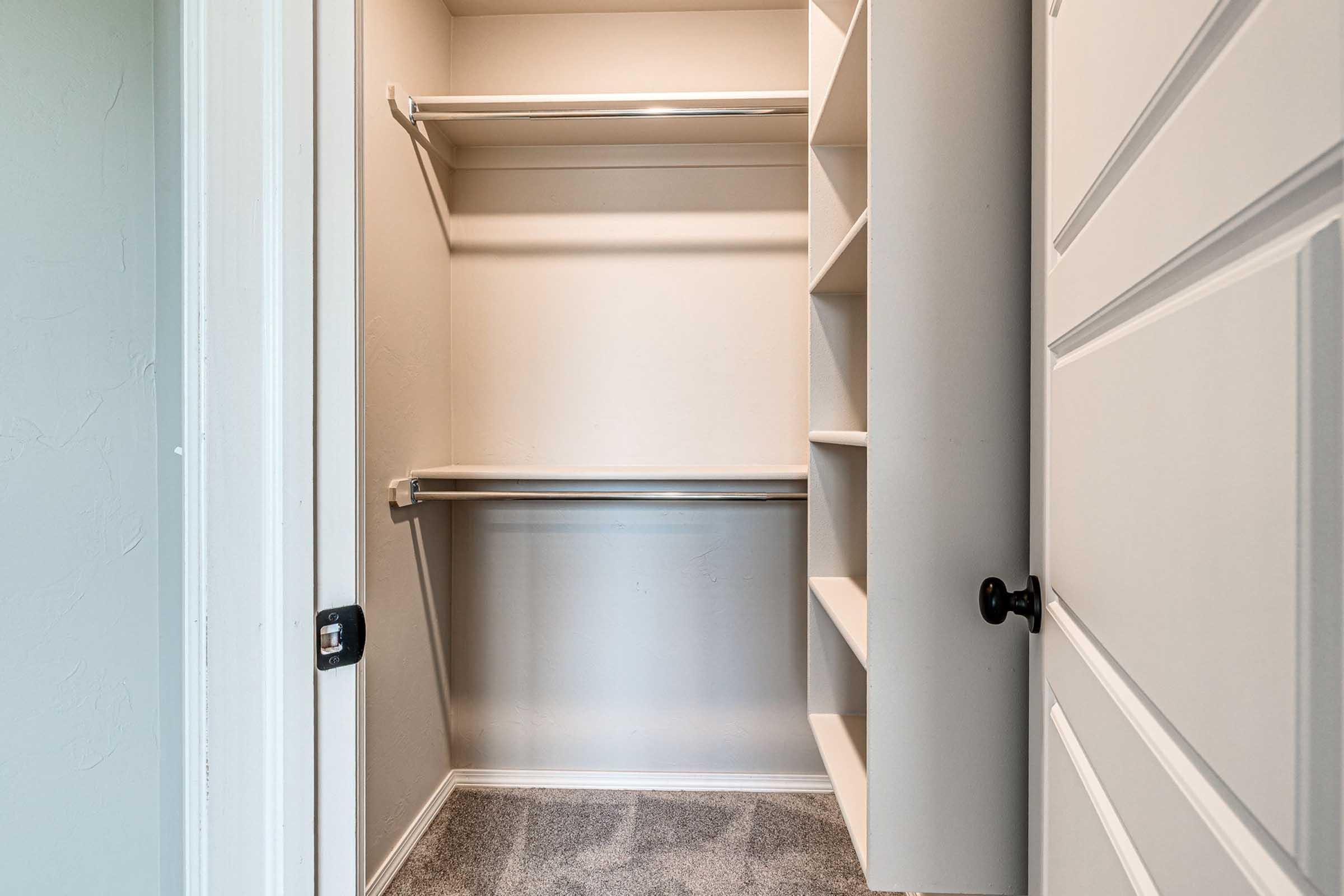 An empty walk-in closet featuring two shelves on the back wall, a side shelving unit, and carpeted flooring. The closet is painted a light color, and the door has a black handle.