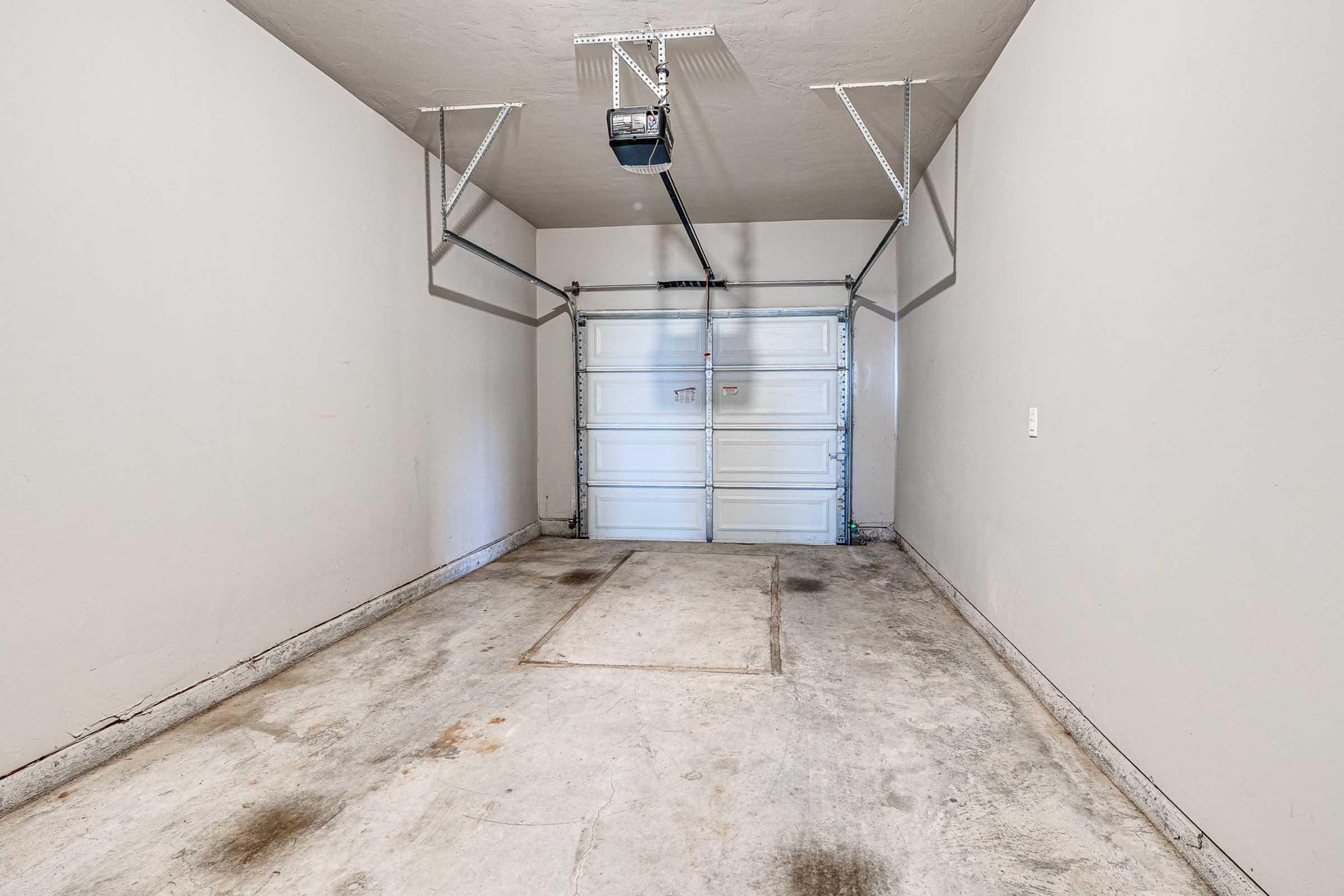 Empty garage with concrete flooring and light gray walls. The garage door is closed, featuring a horizontal panel design. Overhead, a garage door opener is installed. The space is unoccupied and well-lit, giving a clean and minimalistic appearance.