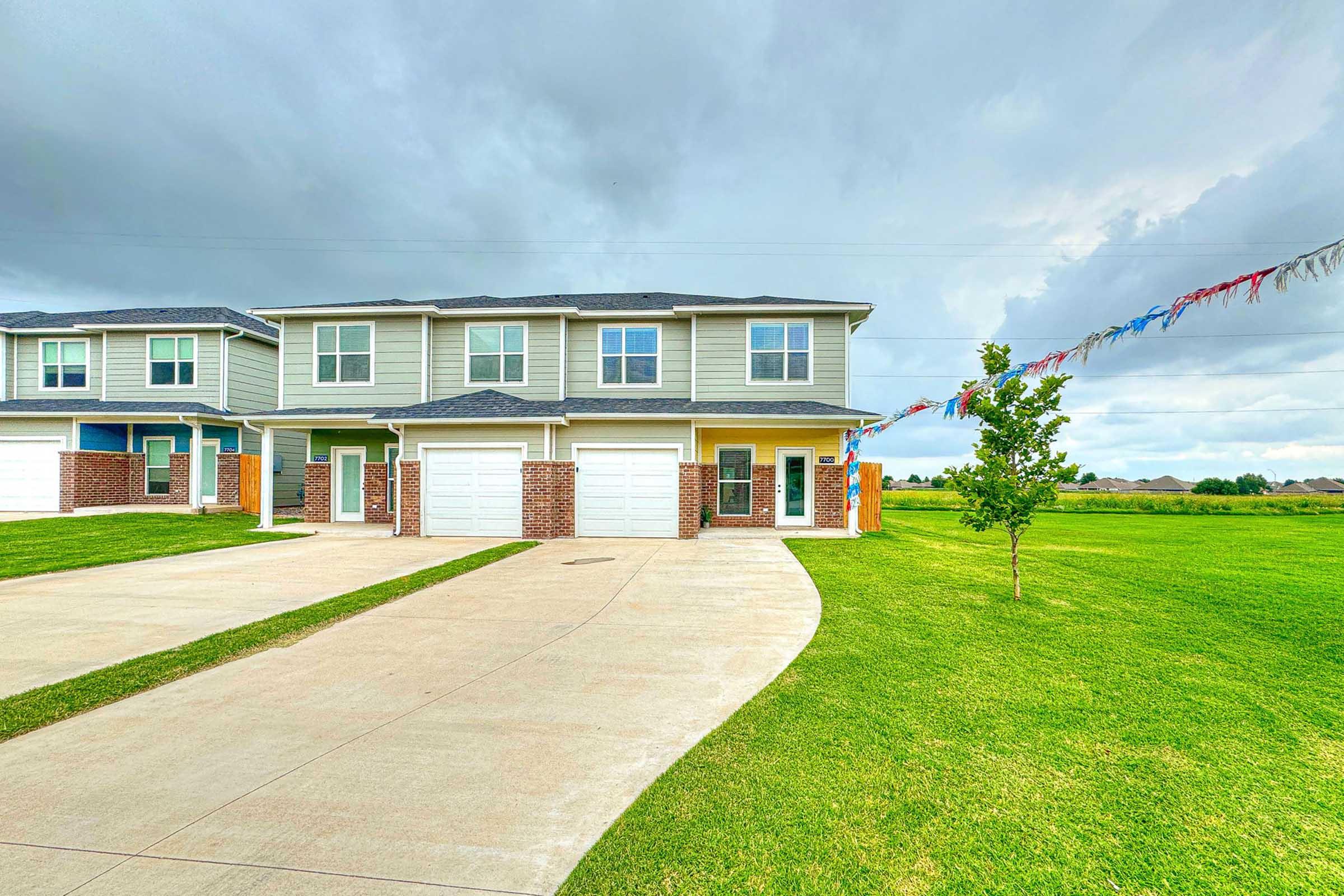A modern two-story duplex with a well-maintained lawn. Each unit has a garage, and colorful flags are strung along the driveway. The sky is partly cloudy, and surrounding fields can be seen in the background.